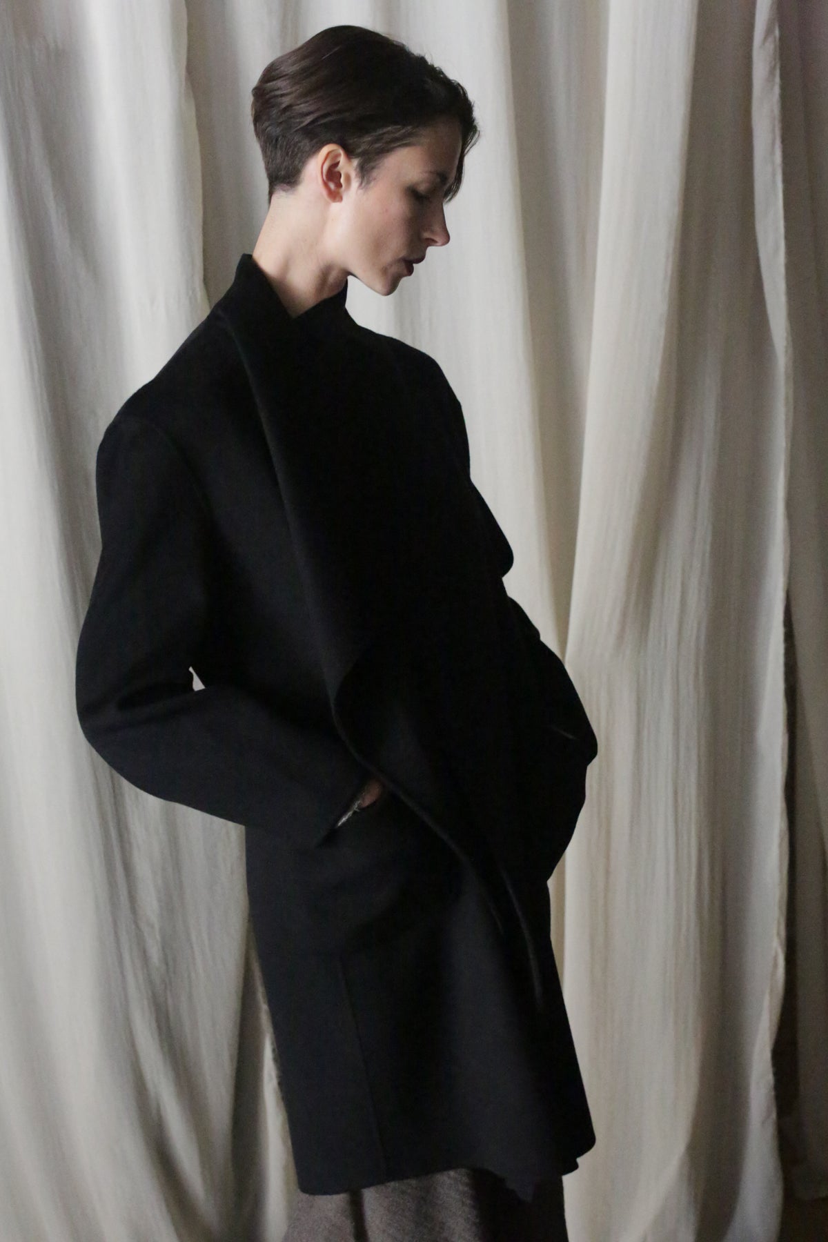 A person with short hair stands in profile wearing the Winston Long Double-face coat in black, hands in pockets, against draped white fabric. Soft lighting enhances the dramatic classic hues of this wardrobe foundation piece.