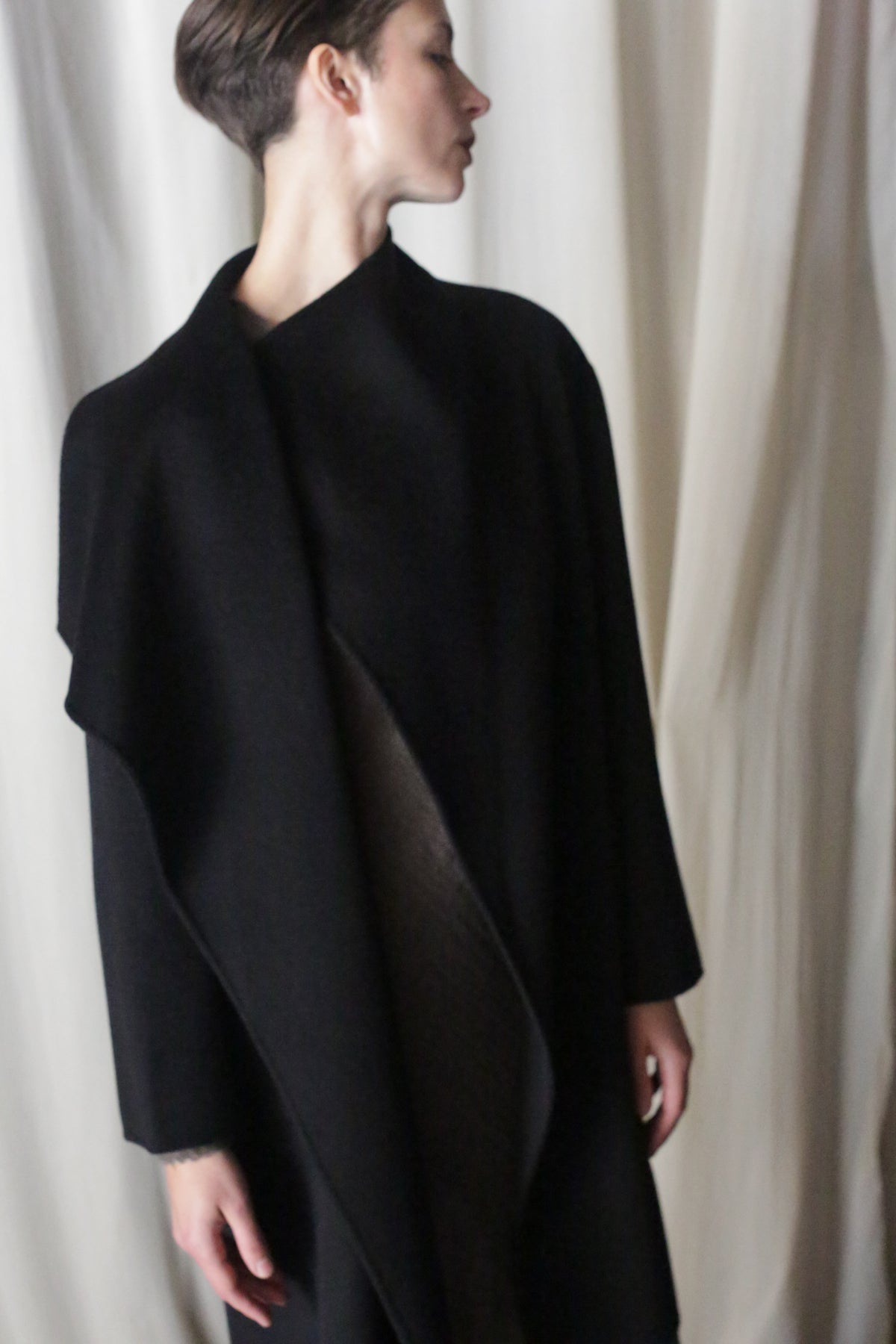 Wearing the Winston Long Double-face in Black, someone with short hair stands sideways against softly draped fabric, gazing upward. This timeless piece is a wardrobe essential for any style enthusiast.