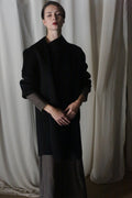 The person models the Winston Long Double-face coat in black over a brown dress, epitomizing classic wardrobe essentials. With hands clasped against a backdrop of gentle white drapery, the scene exudes simplicity and elegance.