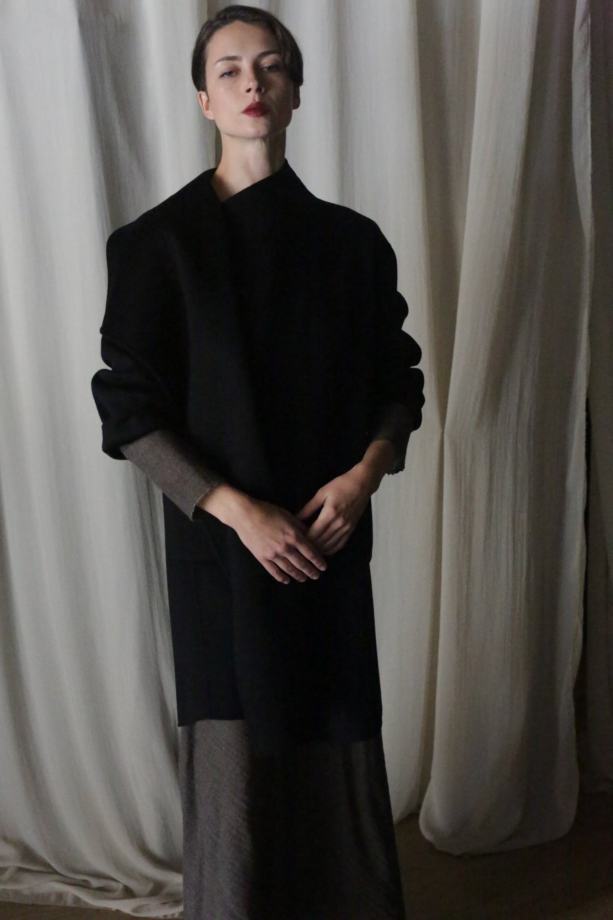 The person models the Winston Long Double-face coat in black over a brown dress, epitomizing classic wardrobe essentials. With hands clasped against a backdrop of gentle white drapery, the scene exudes simplicity and elegance.