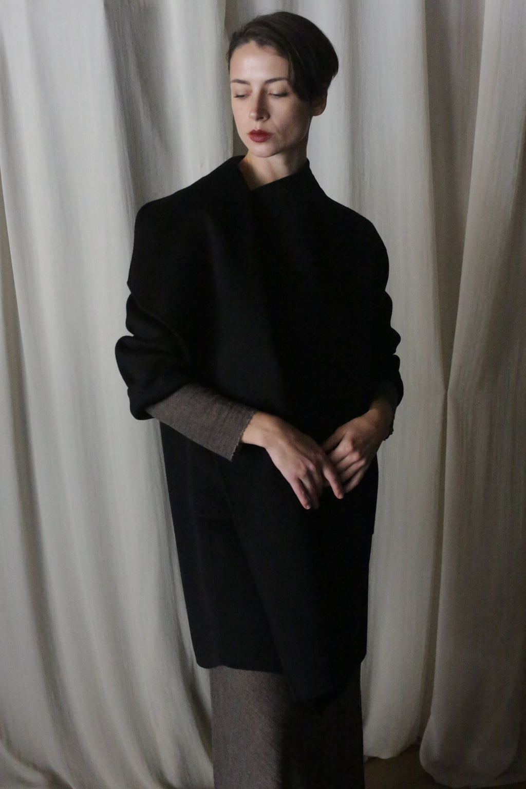 A person stands before draped white curtains, wearing the Winston Long Double-face coat in black over lighter clothing. They have short, styled hair and a poised expression, with hands gently clasped. The classic hues and dim lighting create a serene, contemplative mood.