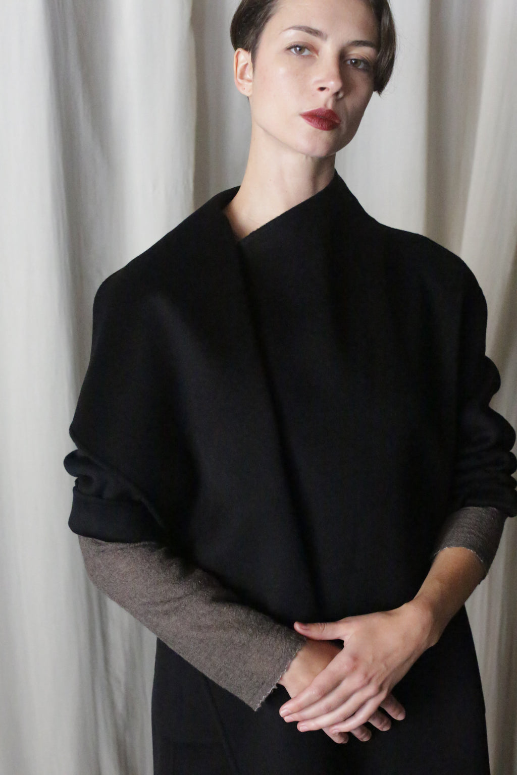 Standing against a light curtain in the Winston Long Double-face | Black, a person with short hair embodies timeless elegance. Crafted from double-faced cashmere and draped elegantly, they look slightly upward with hands clasped, making it a perfect piece for any wardrobe foundation.