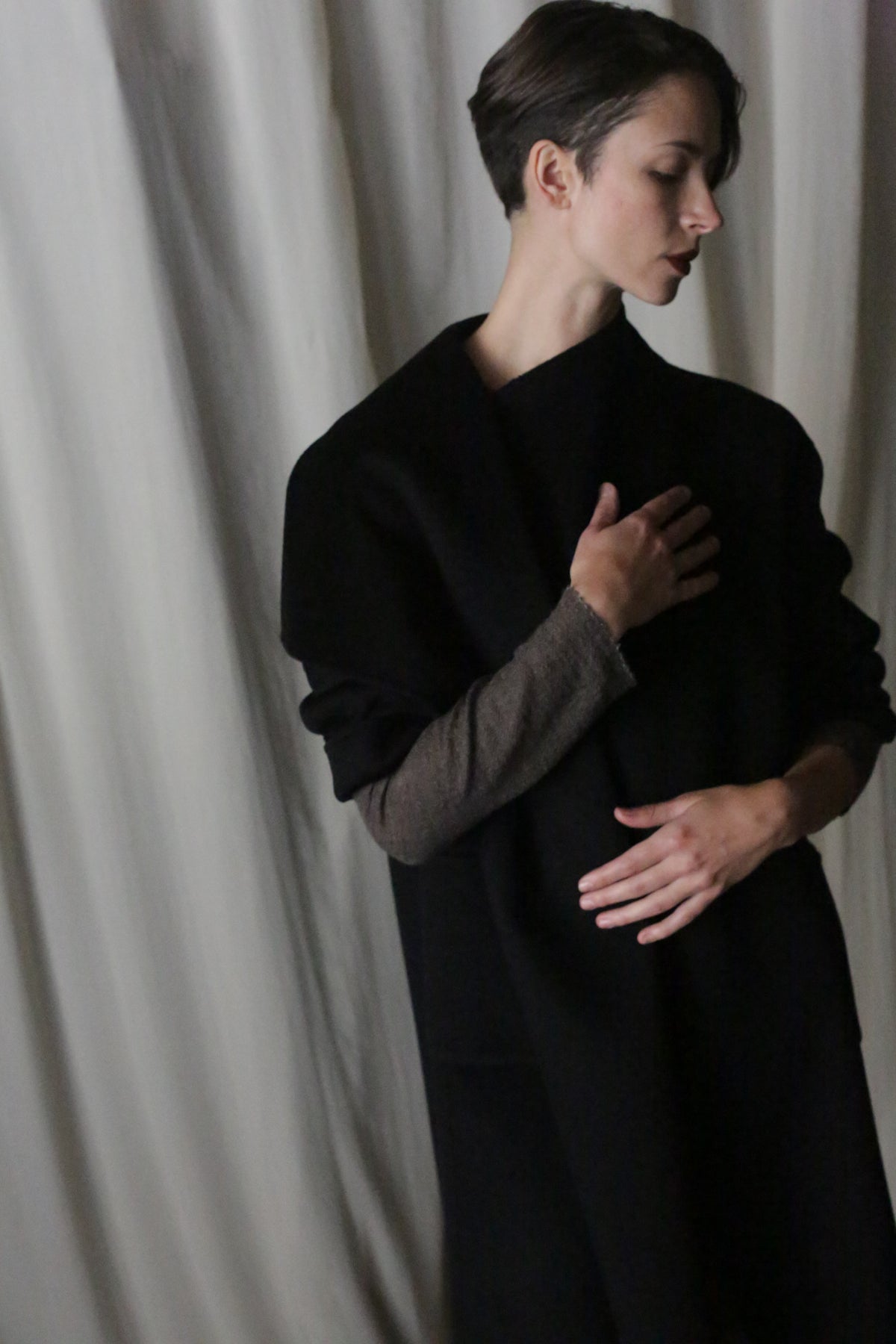 A person with short hair stands gracefully against draped white fabric, wearing the Winston Long Double-face in Black. They have a contemplative expression, one hand gently on their chest while the other rests by their side.