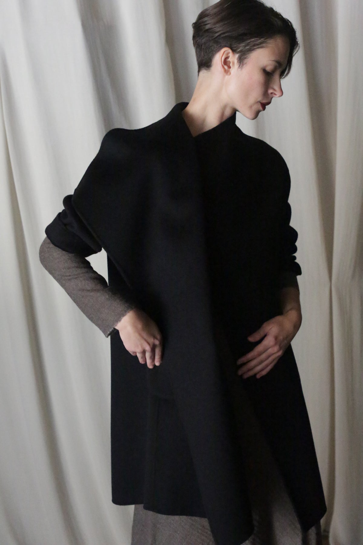 A person with short hair poses in front of a light curtain, wearing the stylish Winston Long Double-face coat in black over a taupe outfit. They stand slightly turned, one hand on their chest and the other on their waist, highlighting classic hues as essential wardrobe staples.
