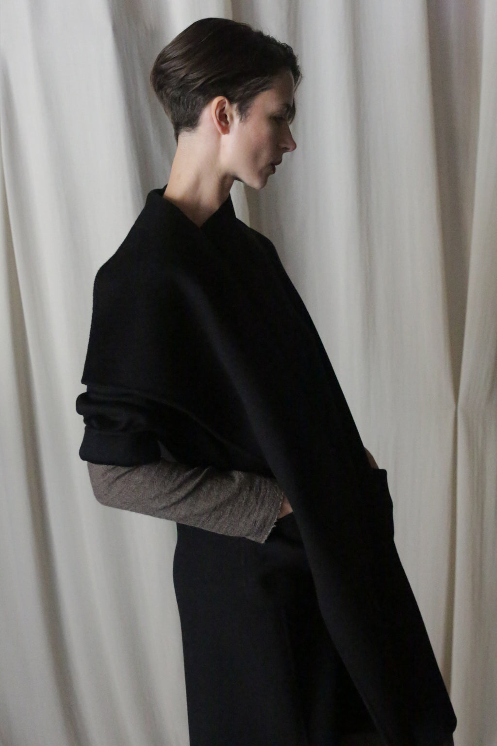 A person with short hair stands in profile against a cream-colored curtain, draped in the Winston Long Double-face | Black shawl made of double-faced cashmere over a light long-sleeve top. The soft lighting creates shadows for a minimalist and contemplative feel.