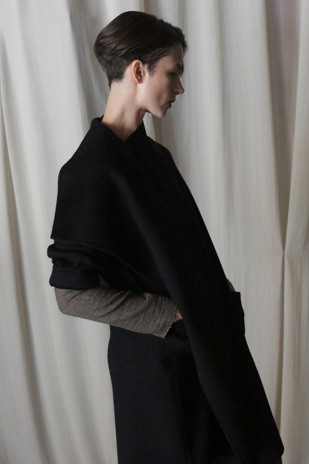 A person with short hair stands in profile against a cream-colored curtain, draped in the Winston Long Double-face | Black shawl made of double-faced cashmere over a light long-sleeve top. The soft lighting creates shadows for a minimalist and contemplative feel.