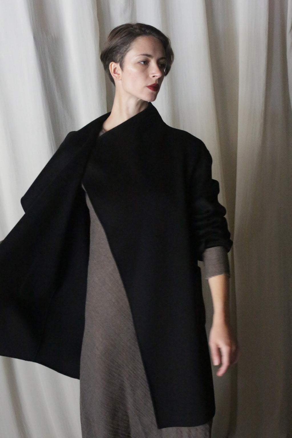 A person with short hair, wearing the Winston Long Double-face in Black over a textured, earth-toned dress depicting timeless neutrals, stands before a light draped fabric background. Their gaze is directed to the side, one arm subtly raised.