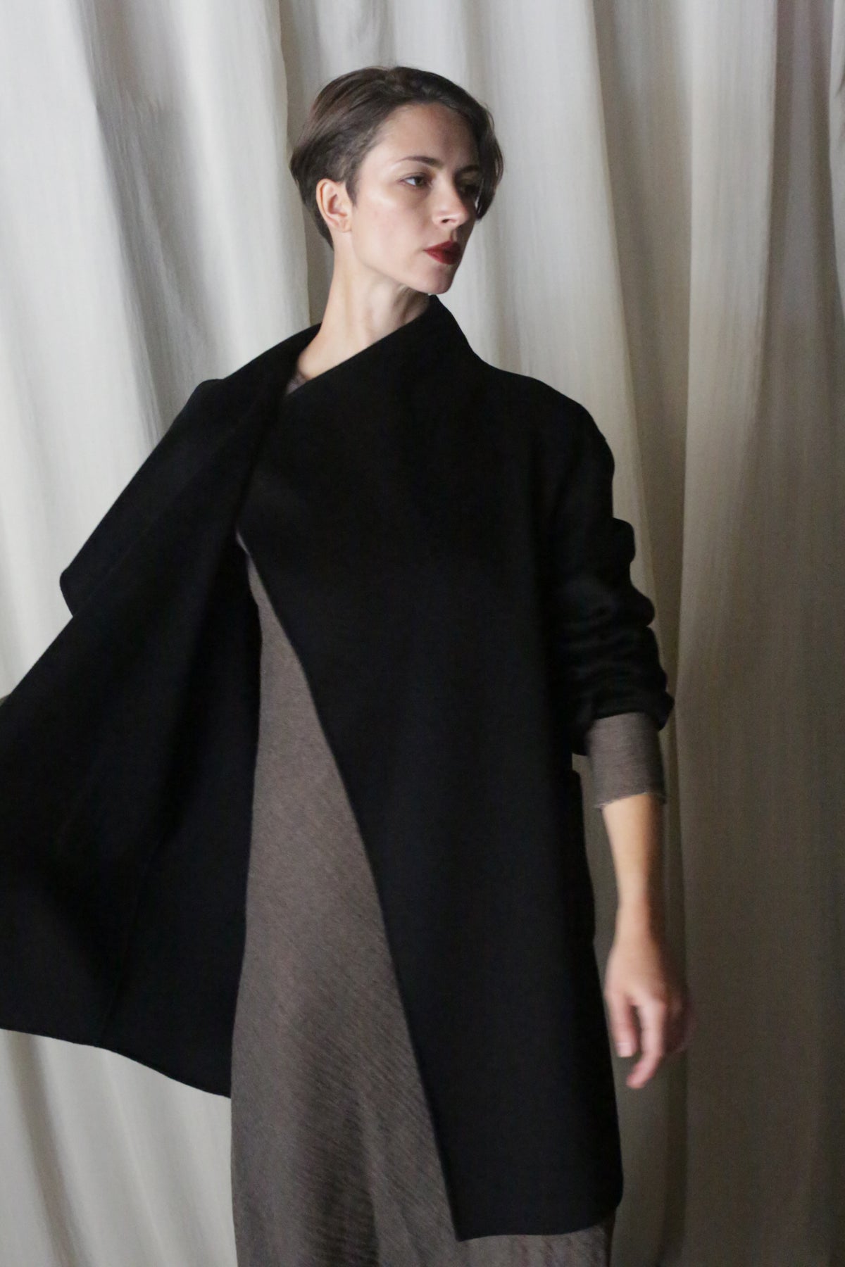 A person with short hair, wearing the Winston Long Double-face in Black over a textured, earth-toned dress depicting timeless neutrals, stands before a light draped fabric background. Their gaze is directed to the side, one arm subtly raised.