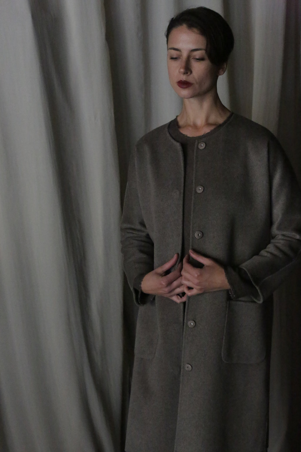 A woman stands with closed eyes and clasped hands, wearing the Battersea Double-Face Cashmere coat in undyed natural. The gray-toned, softly draped fabric background enhances its minimalist aesthetic, creating a serene atmosphere.