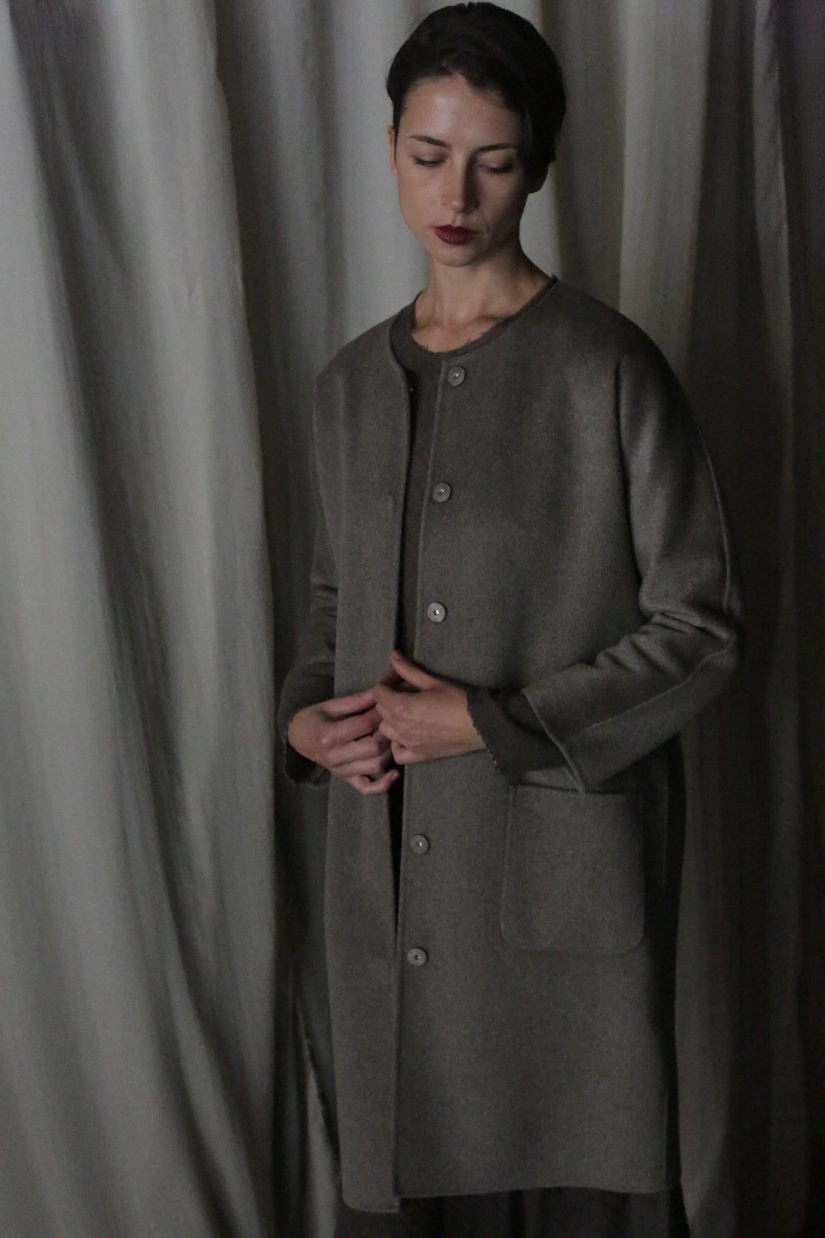 A person with short dark hair stands against draped fabric, wearing the Battersea Double-Face Cashmere coat in undyed natural. This luxurious, long coat has large pockets and a versatile silhouette. They have a serious expression, looking slightly downward with hands gently clasped.