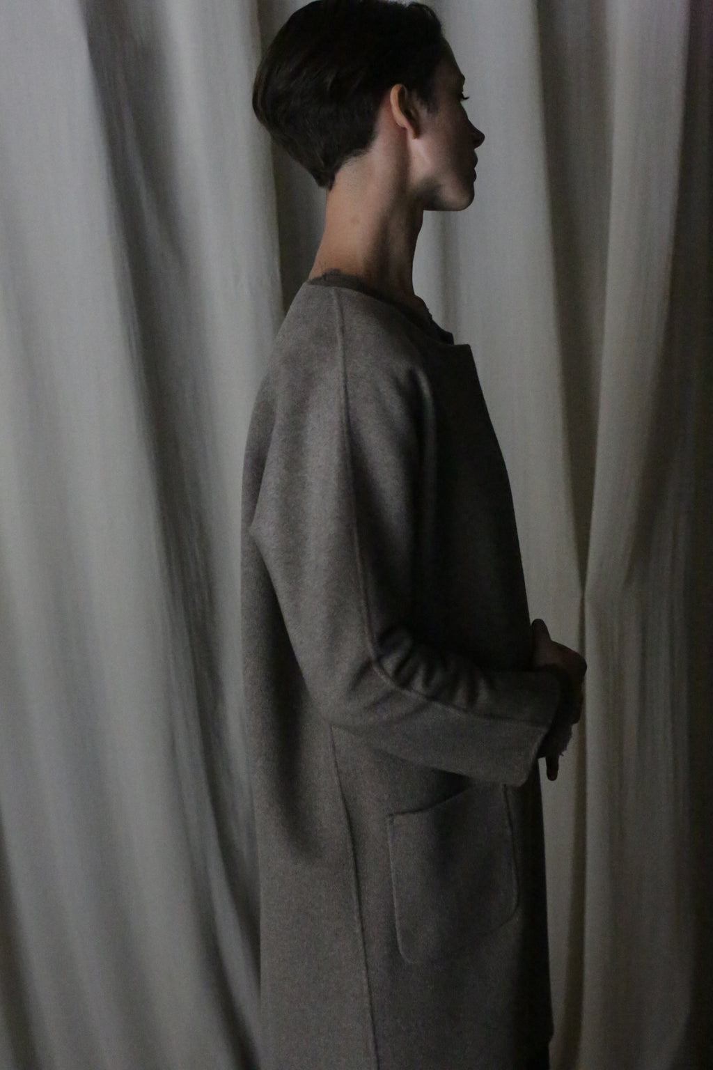 A person with short hair stands in profile, wearing the versatile Battersea Double-Face Cashmere coat in undyed natural, featuring a minimalist silhouette with large pockets. It enhances the aesthetic against a softly lit background with draped fabric.