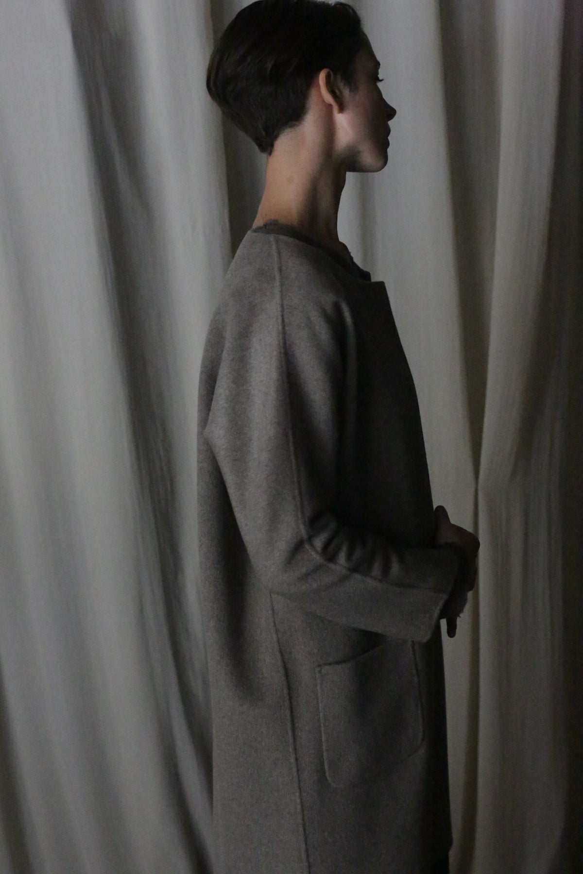 A person with short hair stands in profile, wearing the versatile Battersea Double-Face Cashmere coat in undyed natural, featuring a minimalist silhouette with large pockets. It enhances the aesthetic against a softly lit background with draped fabric.