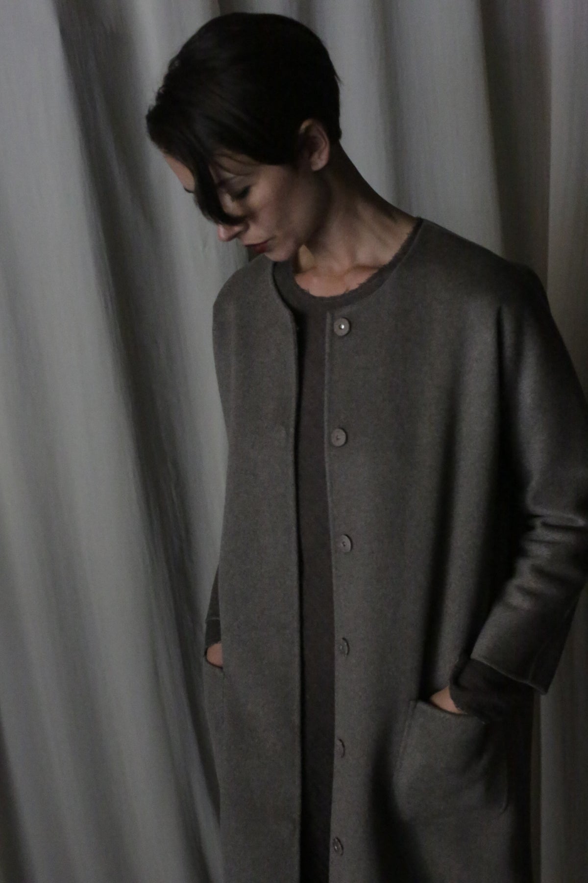 A person with short dark hair stands against a curtain, looking down. They wear the Battersea Double-Face Cashmere coat in undyed natural, hands tucked into the pockets. Soft lighting creates a subdued atmosphere.