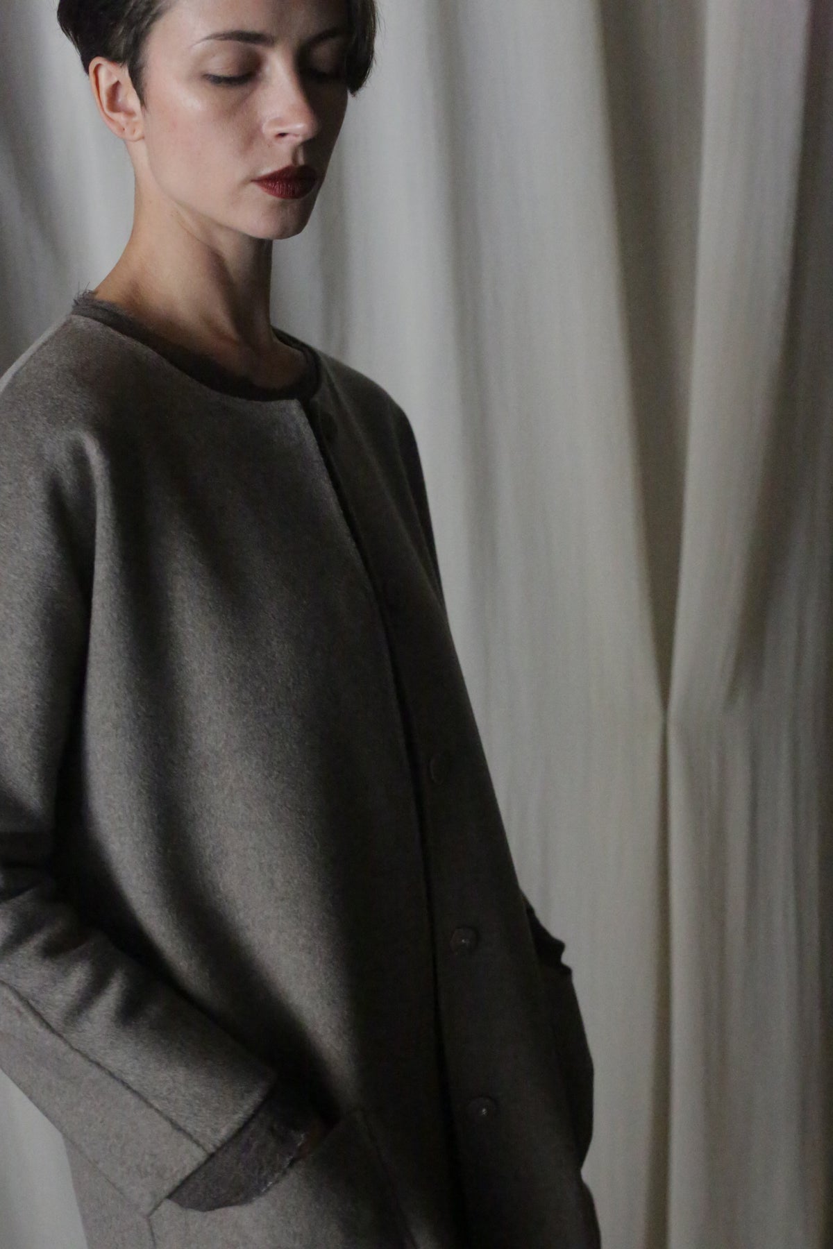 A person with short dark hair stands with eyes closed, wrapped in the Battersea Double-Face Cashmere coat in undyed natural. The softly draped fabric in neutral tones serves as the perfect backdrop for this versatile silhouette.