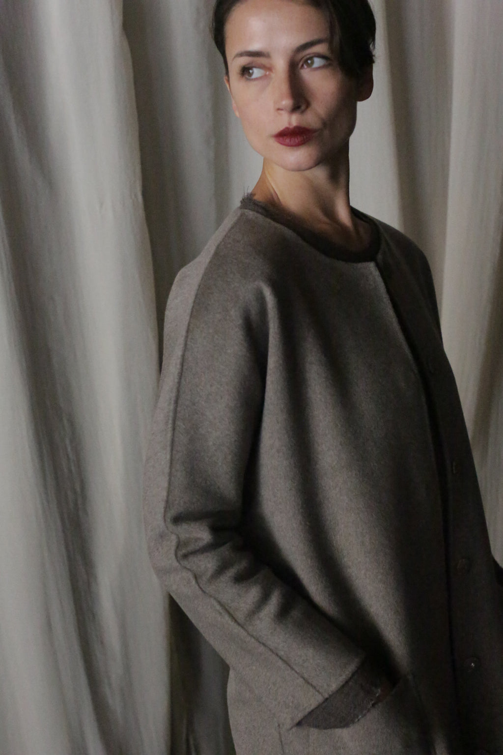 A person with short dark hair stands contemplatively, wearing the Battersea Double-Face Cashmere in Undyed Natural. The coat's versatile silhouette and side pockets create a minimalist aesthetic complemented by light-colored curtains in the background.