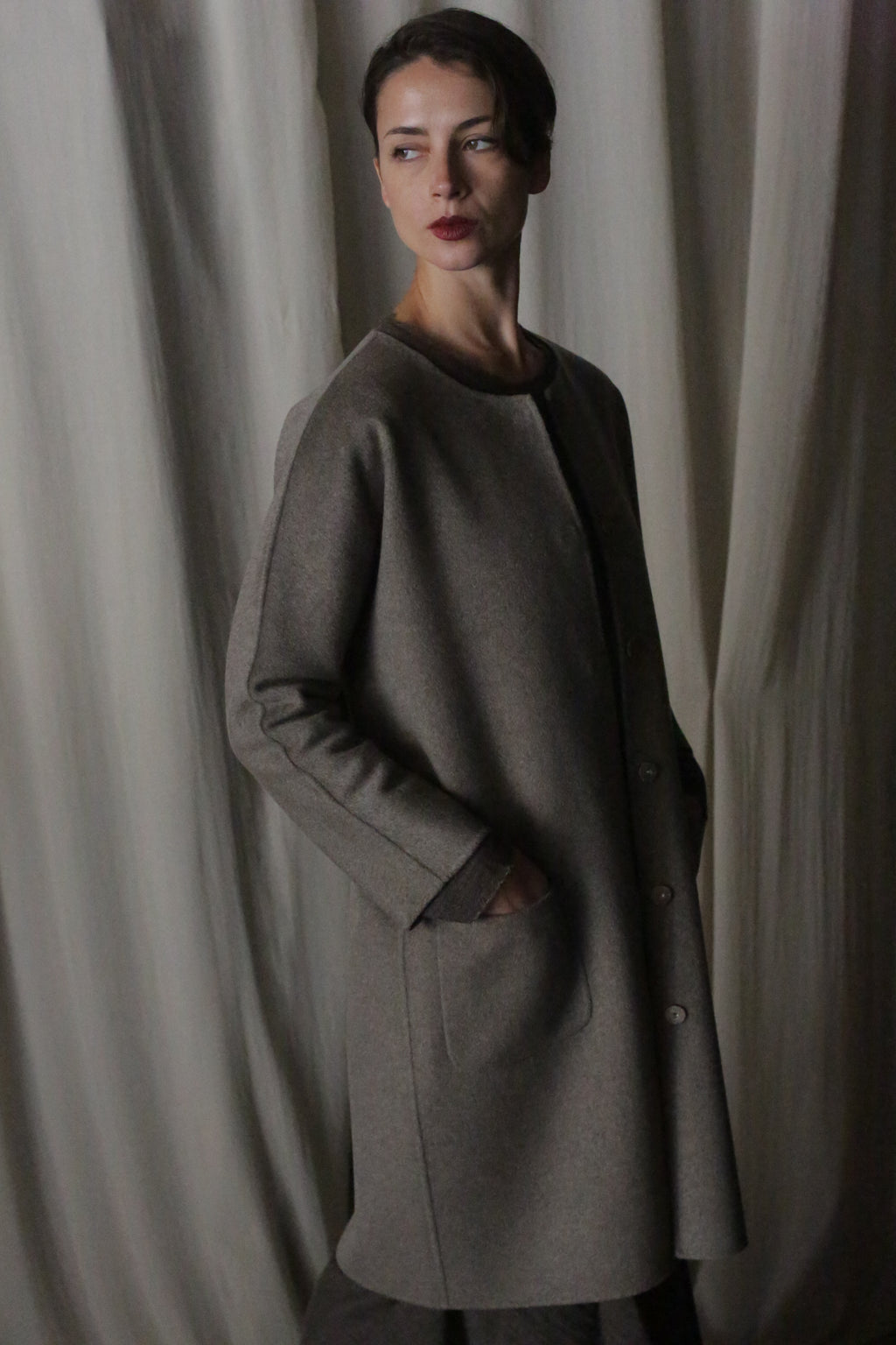 Against a softly draped fabric backdrop, a person wears the Battersea Double-Face Cashmere coat in undyed natural, exuding luxury. With hands in pockets and a contemplative look, they stand in soft lighting that enhances the coat's versatile silhouette and creates an elegant atmosphere.