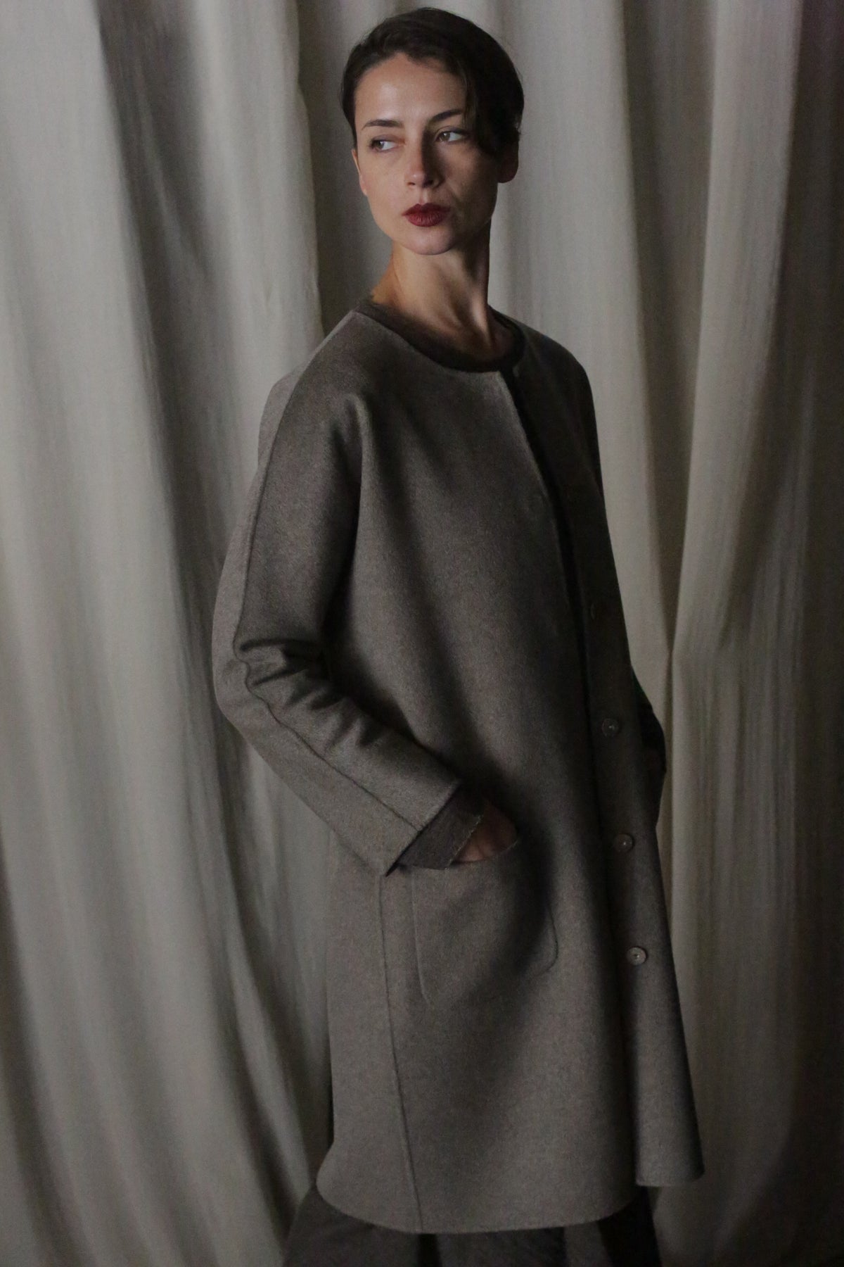 Against a softly draped fabric backdrop, a person wears the Battersea Double-Face Cashmere coat in undyed natural, exuding luxury. With hands in pockets and a contemplative look, they stand in soft lighting that enhances the coat's versatile silhouette and creates an elegant atmosphere.