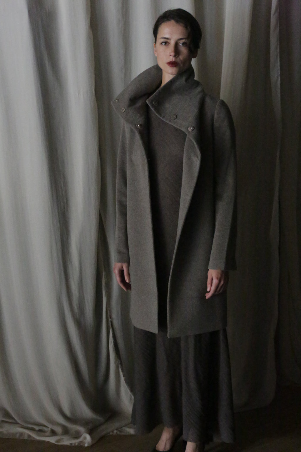 A person stands before draped fabric in a dark, high-collared cashmere coat and Stamford Long | Undyed Natural dress. Dim lighting sets a moody, elegant scene reminiscent of quiet evenings in Stamford.