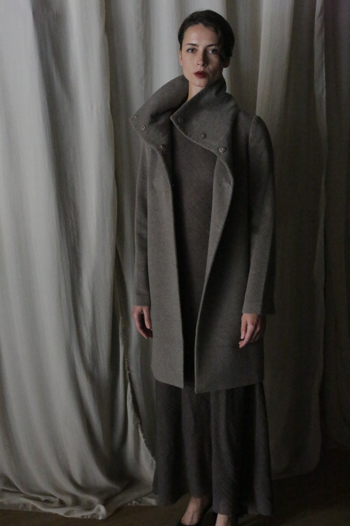 A person stands before draped fabric in a dark, high-collared cashmere coat and Stamford Long | Undyed Natural dress. Dim lighting sets a moody, elegant scene reminiscent of quiet evenings in Stamford.