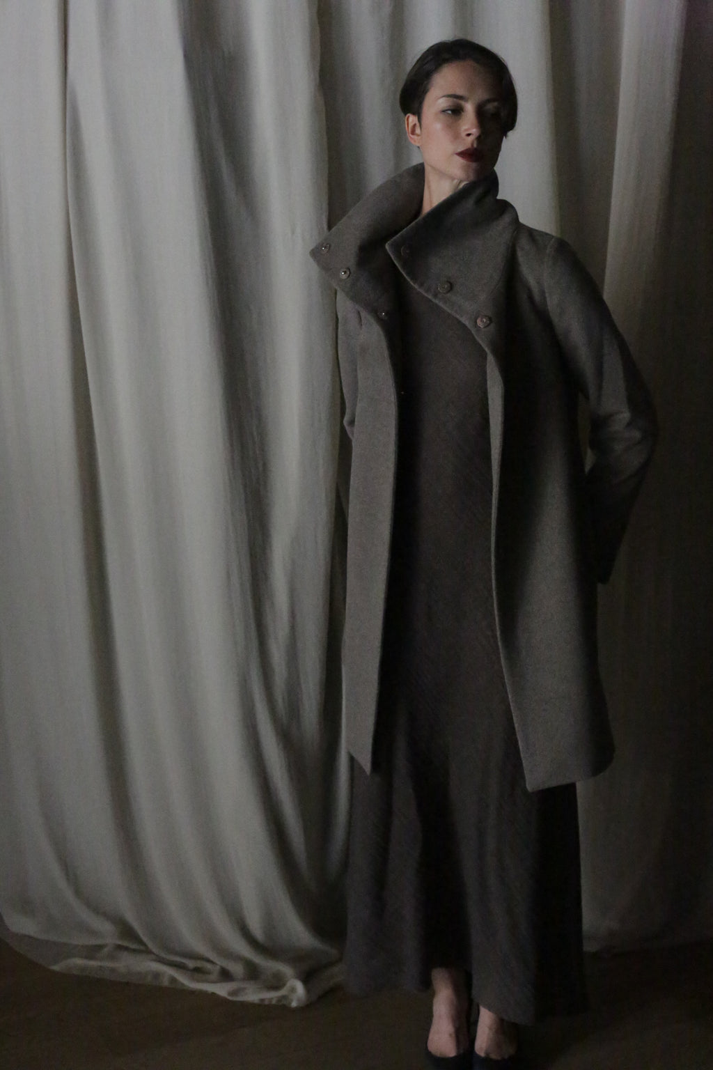 In front of a softly draped curtain, a person wears the Stamford Long | Undyed Natural cashmere coat with a high collar and matching dress. The dim lighting casts shadows, creating an elegant, moody atmosphere inspired by Stamford.