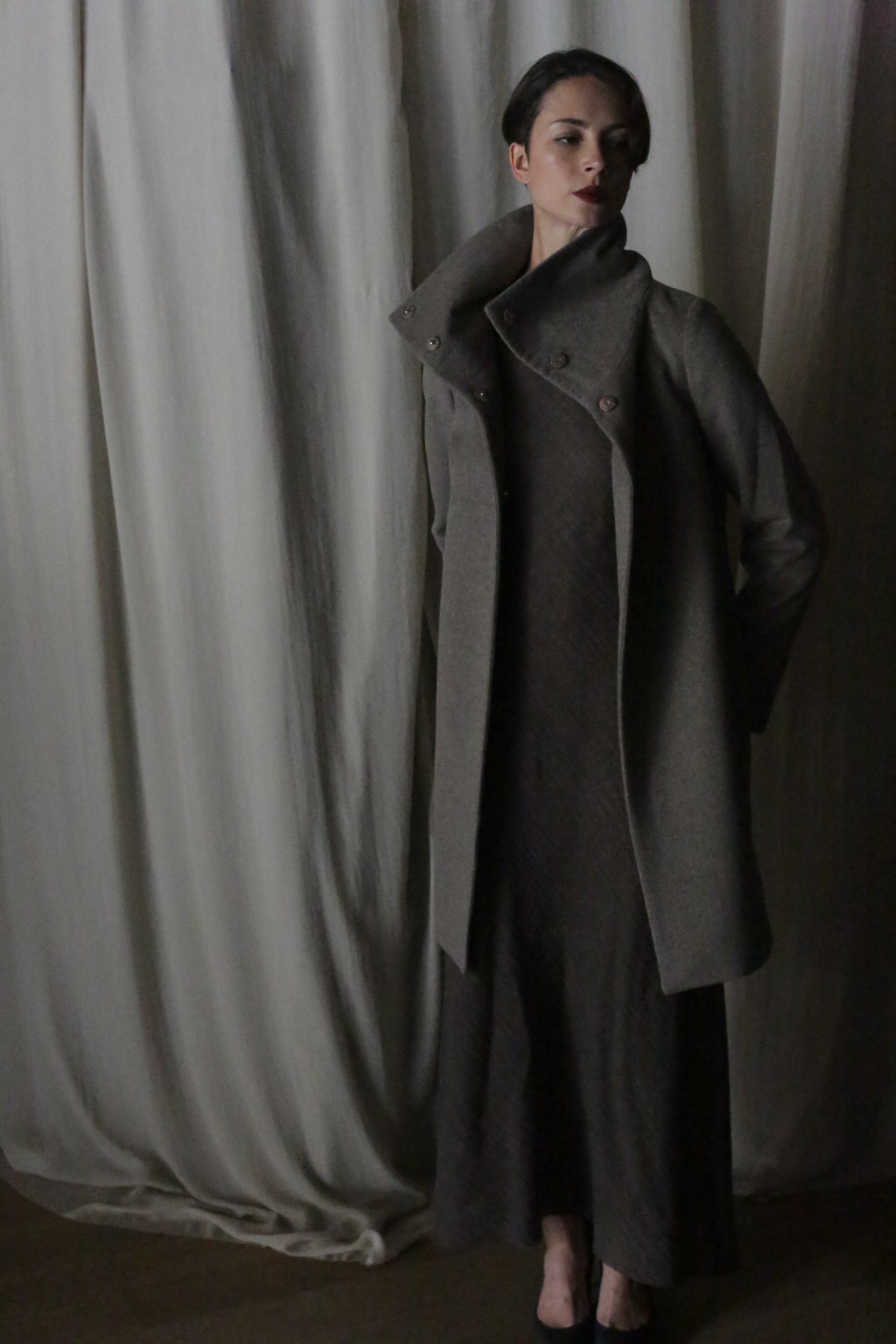 In front of a softly draped curtain, a person wears the Stamford Long | Undyed Natural cashmere coat with a high collar and matching dress. The dim lighting casts shadows, creating an elegant, moody atmosphere inspired by Stamford.
