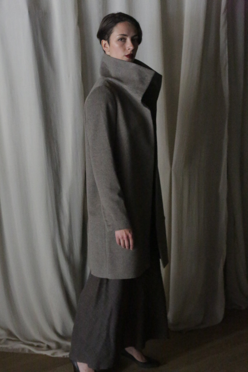 In dim lighting with flowing curtains in the background, a person elegantly poses in profile wearing the Stamford Long coat in Undyed Natural and a dark skirt, gazing towards the camera.