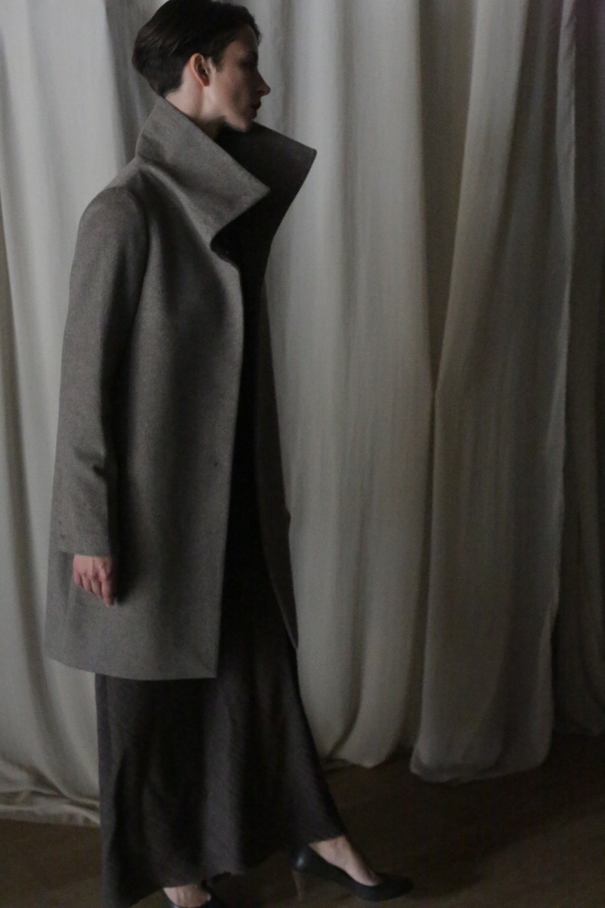 A person in a Stamford Long Undyed Natural cashmere overcoat and dress stands in profile against draped light curtains, their neat short hair complementing the dimly lit, somber elegance. They pair the look with dark shoes.