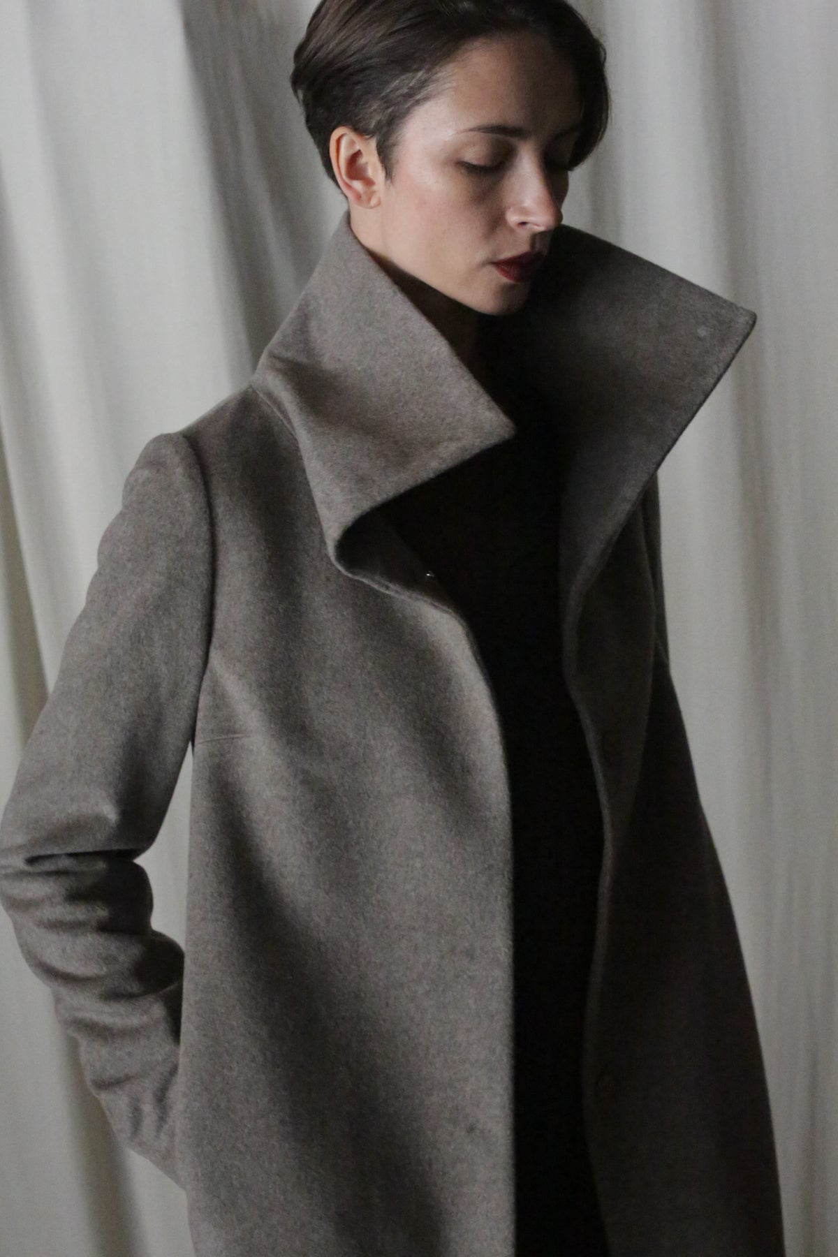 A woman poses serenely in a high-collared Stamford Long | Undyed Natural cashmere coat, hands in pockets. A light curtain serves as a soft, neutral backdrop while gentle lighting highlights the coat's texture.