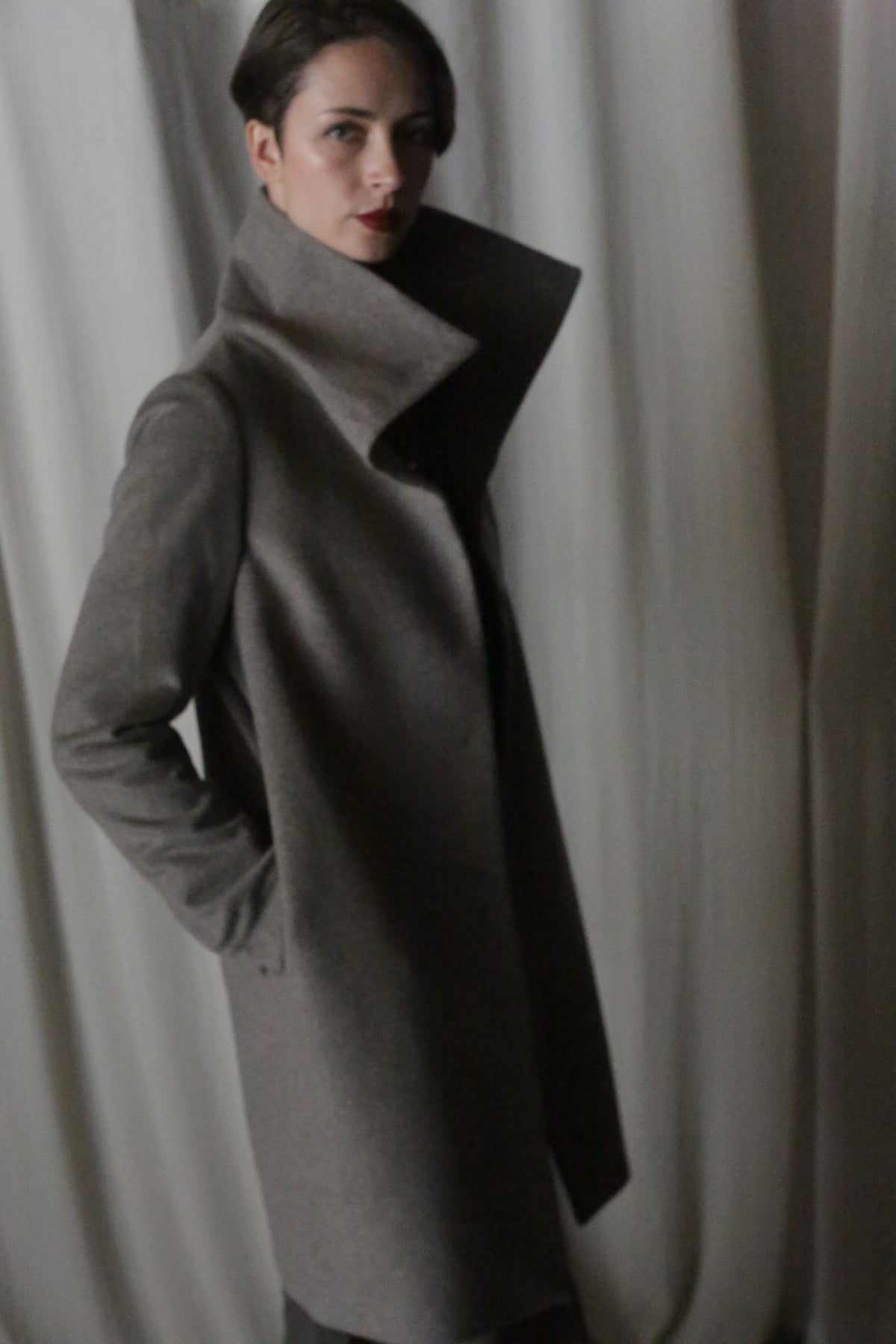 Wearing a Stamford Long coat in Undyed Natural, an individual stands against light fabric with soft shadowed dim lighting, gazing at the camera with a neutral expression—a portrait of timeless elegance.