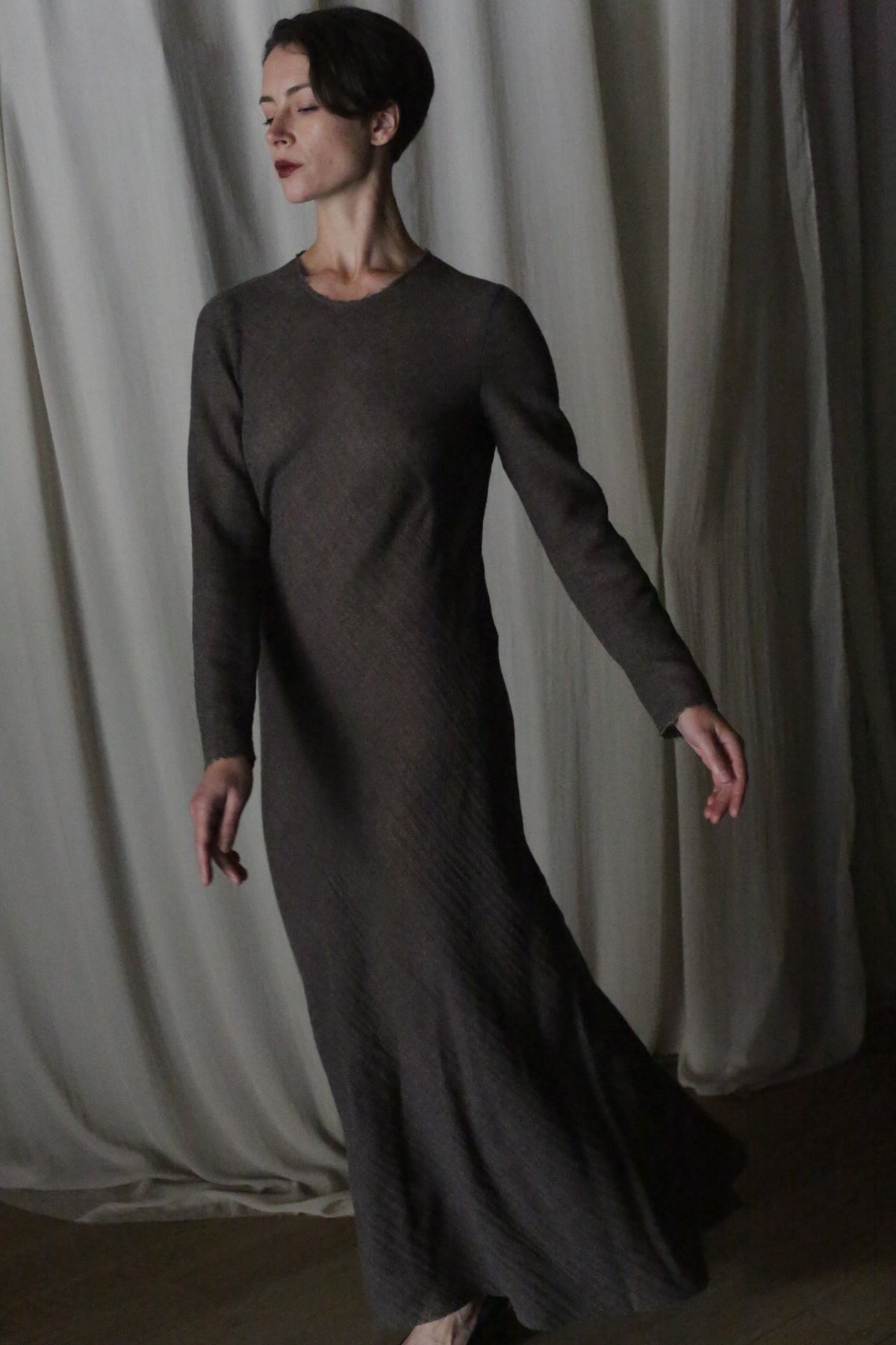 Wearing the Long Sleeved Crewneck Dress, a woman stands with eyes closed and arms gently extended, posed as if dancing against a backdrop of draped, light-colored fabric.