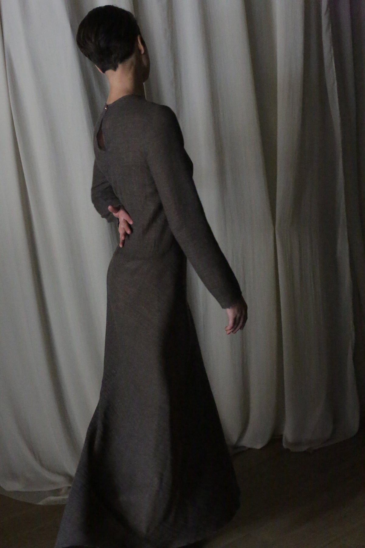 Wearing a Long Sleeved Crewneck Dress, a person stands with their back to the camera and one arm bent, in front of softly draped, light-colored curtains.