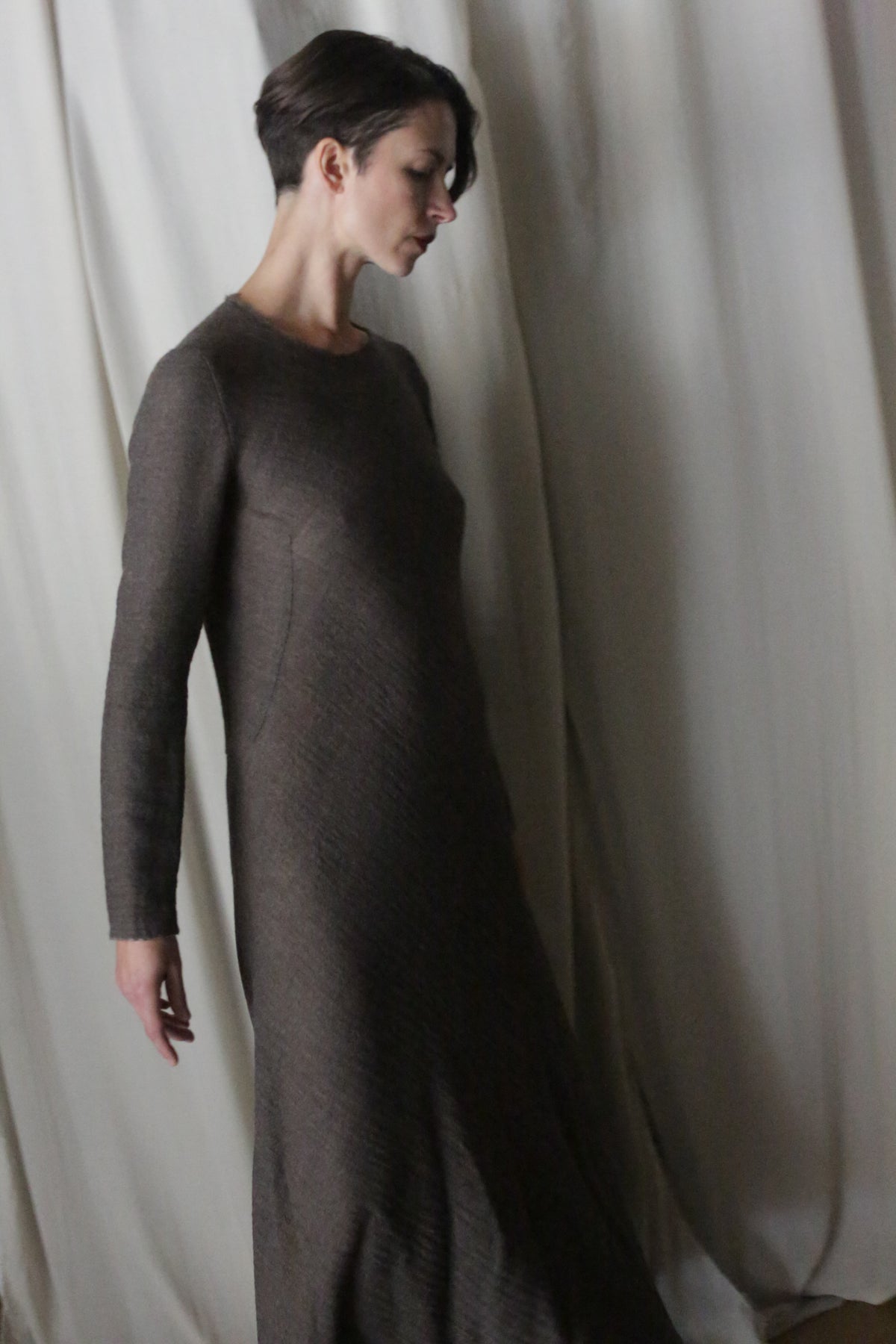 A woman with short dark hair, in a handmade brown Long Sleeved Crewneck Dress, stands before light draped curtains, looking down thoughtfully.