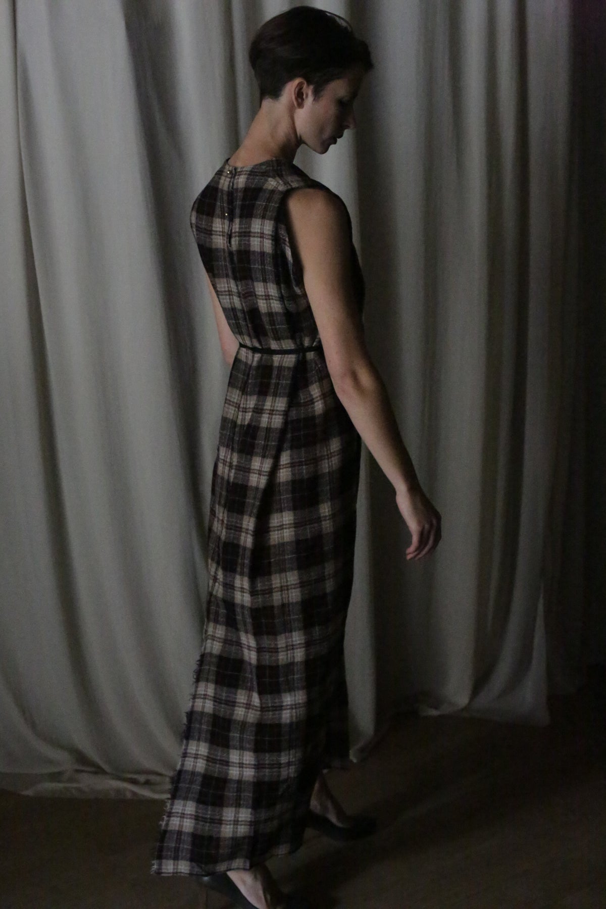 A person with short hair stands sideways in The Winter Dress | Japanese Wool Gauze Taupe Plaid and black heels, posing against draped, light curtains in dim lighting.