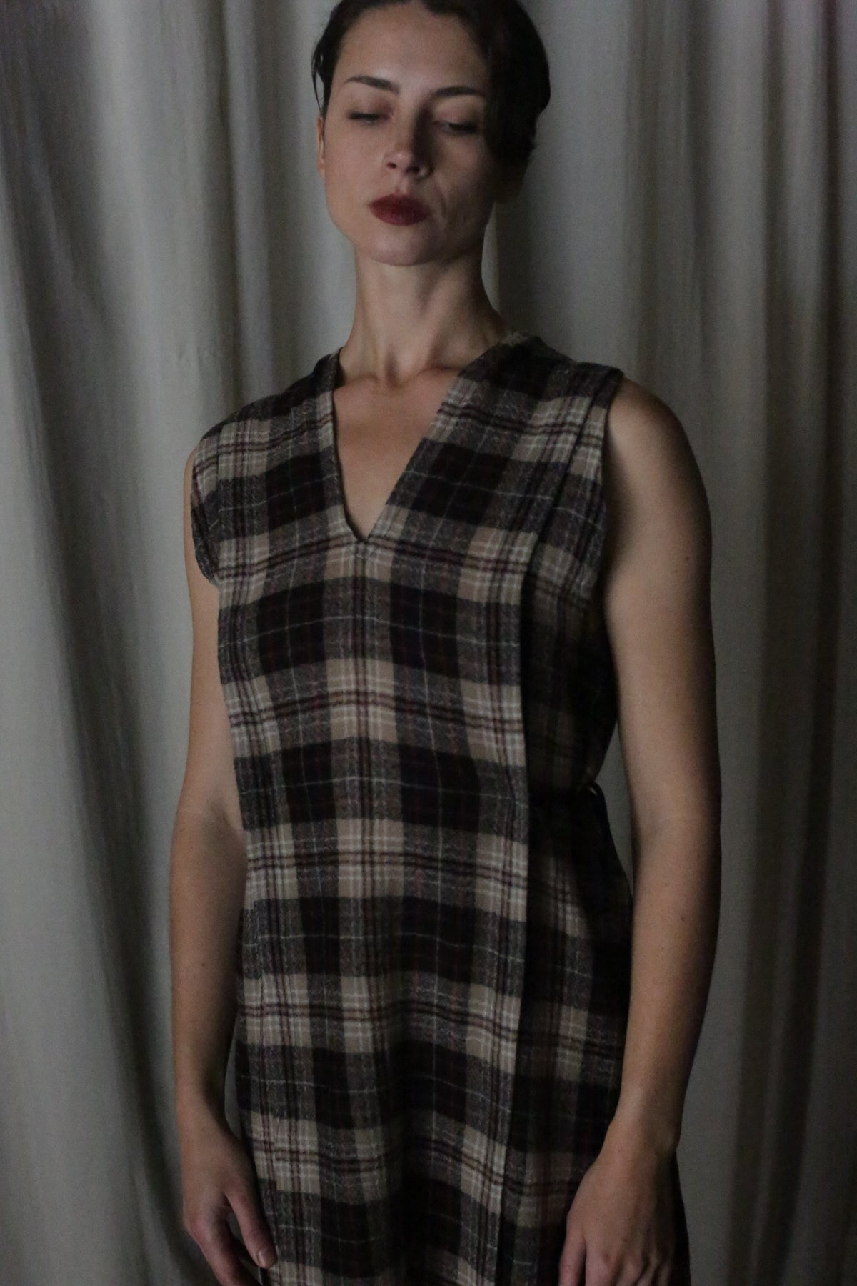 A woman with short dark hair wears The Winter Dress in Japanese Wool Gauze Taupe Plaid, standing before light curtains with her eyes closed and a serene expression.