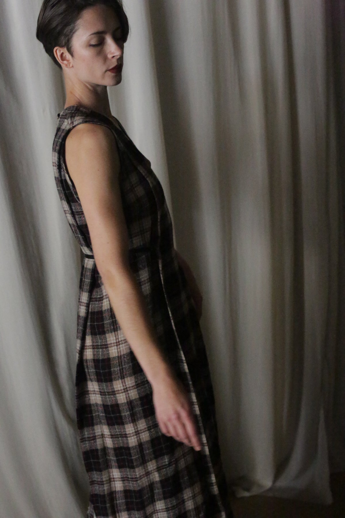A person with short dark hair stands in profile, softly side-lit, wearing The Winter Dress | Japanese Wool Gauze Taupe Plaid against a draped, light-colored fabric backdrop.