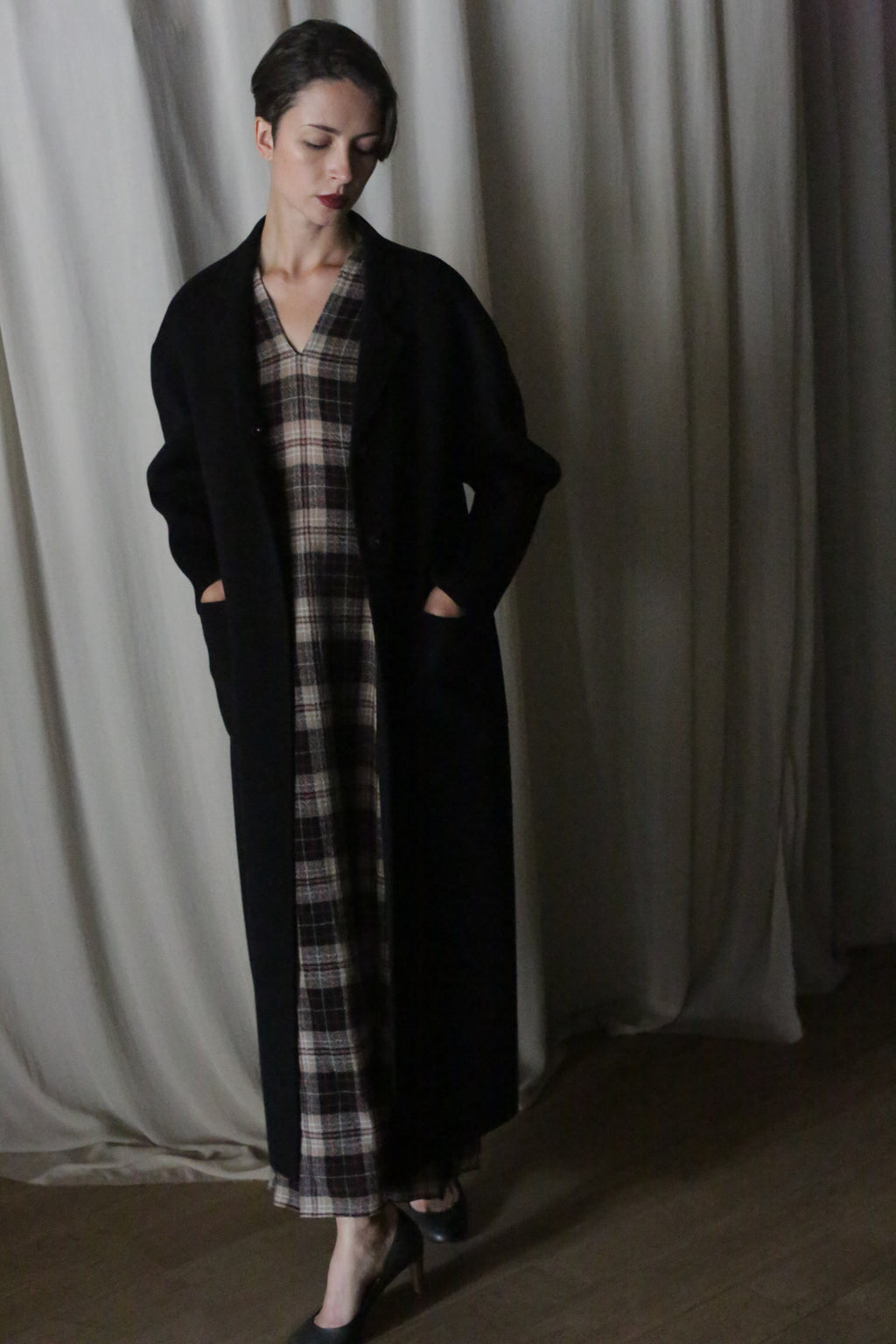 A woman with short hair stands indoors, hands in pockets, wearing the Bamford Long Double-face Cashmere coat in black over a plaid dress and black heels. The backdrop is draped light-colored fabric.