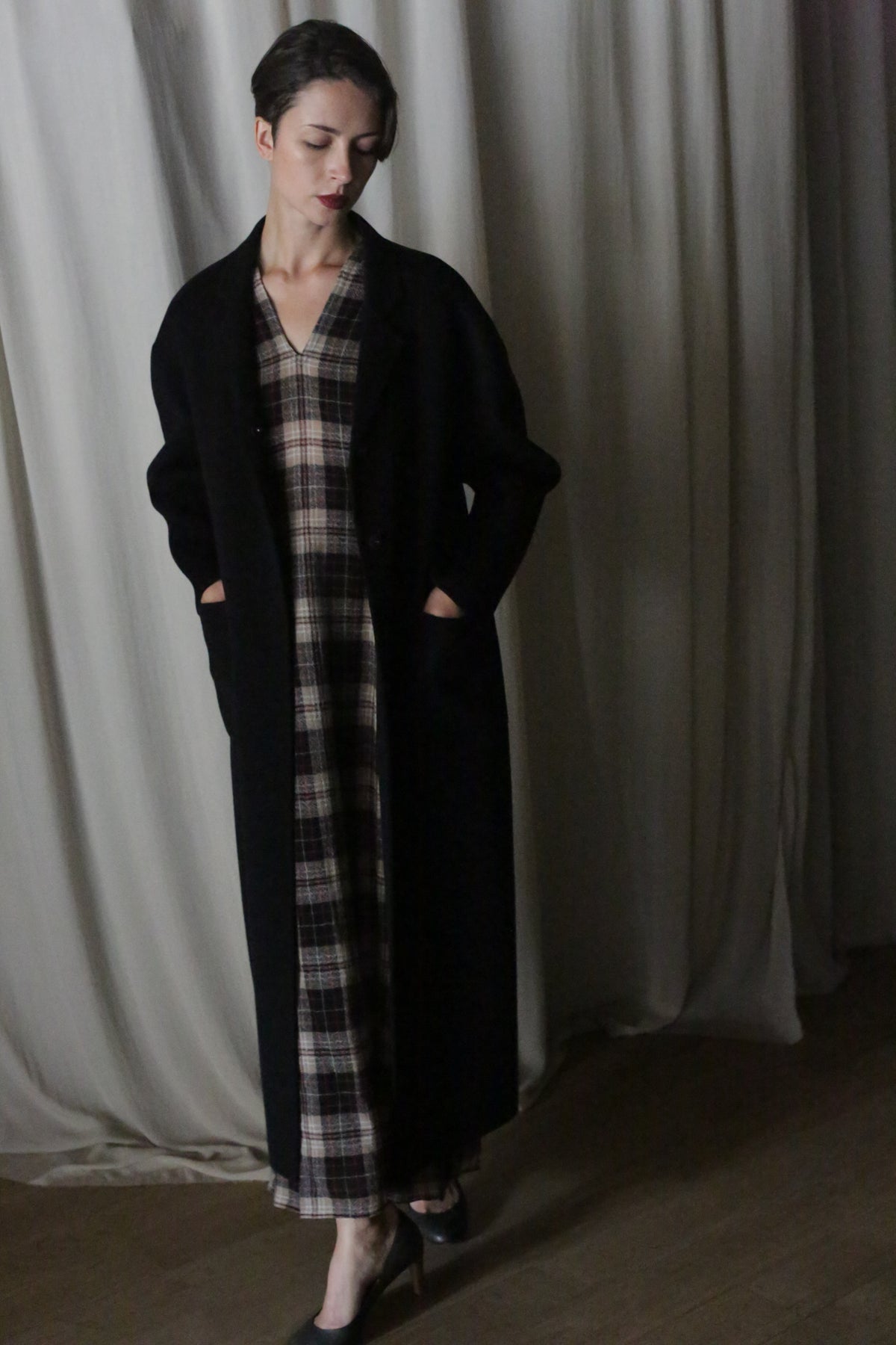 A woman with short hair stands indoors, hands in pockets, wearing the Bamford Long Double-face Cashmere coat in black over a plaid dress and black heels. The backdrop is draped light-colored fabric.