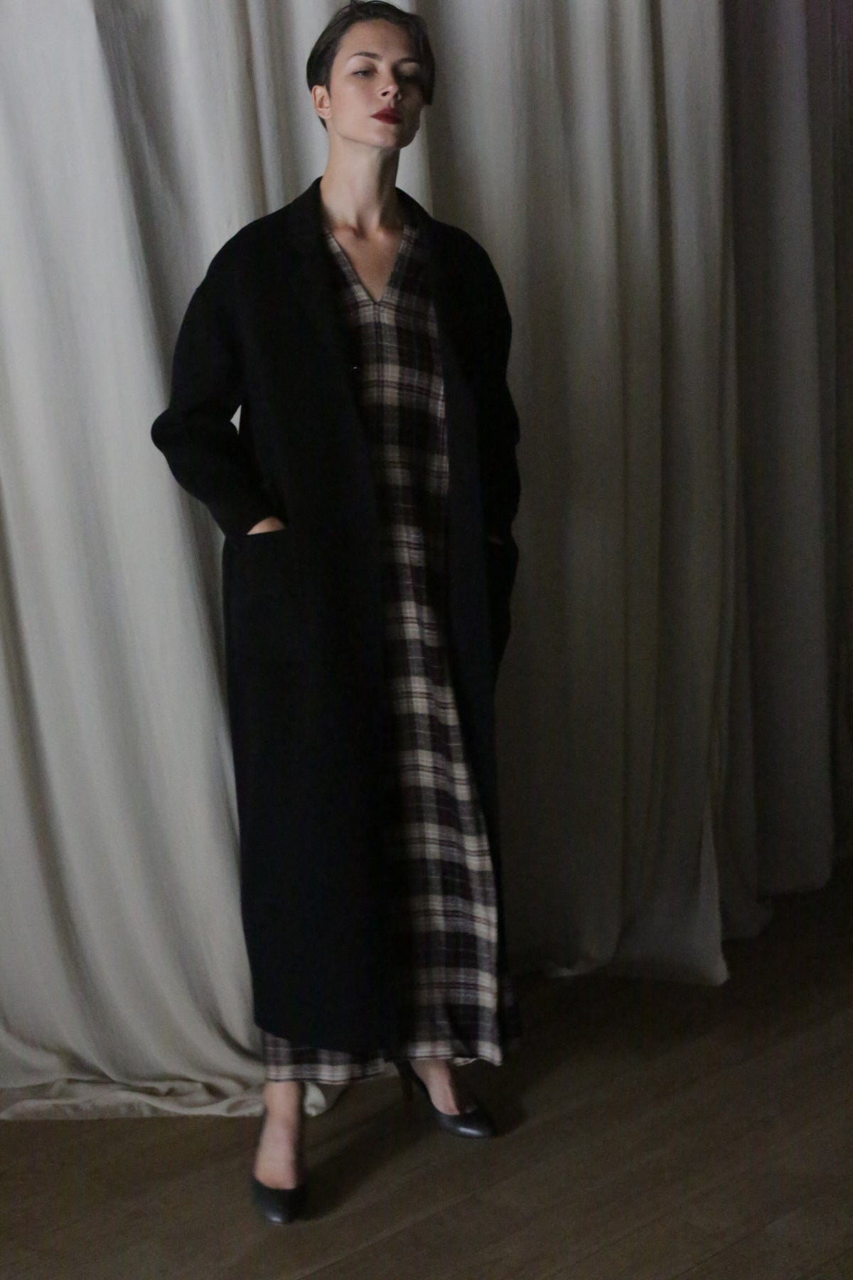 A person stands indoors against light curtains, wearing the Bamford Long Double-face Cashmere coat in black over a plaid dress with dark shoes, hands in pockets and a neutral expression.