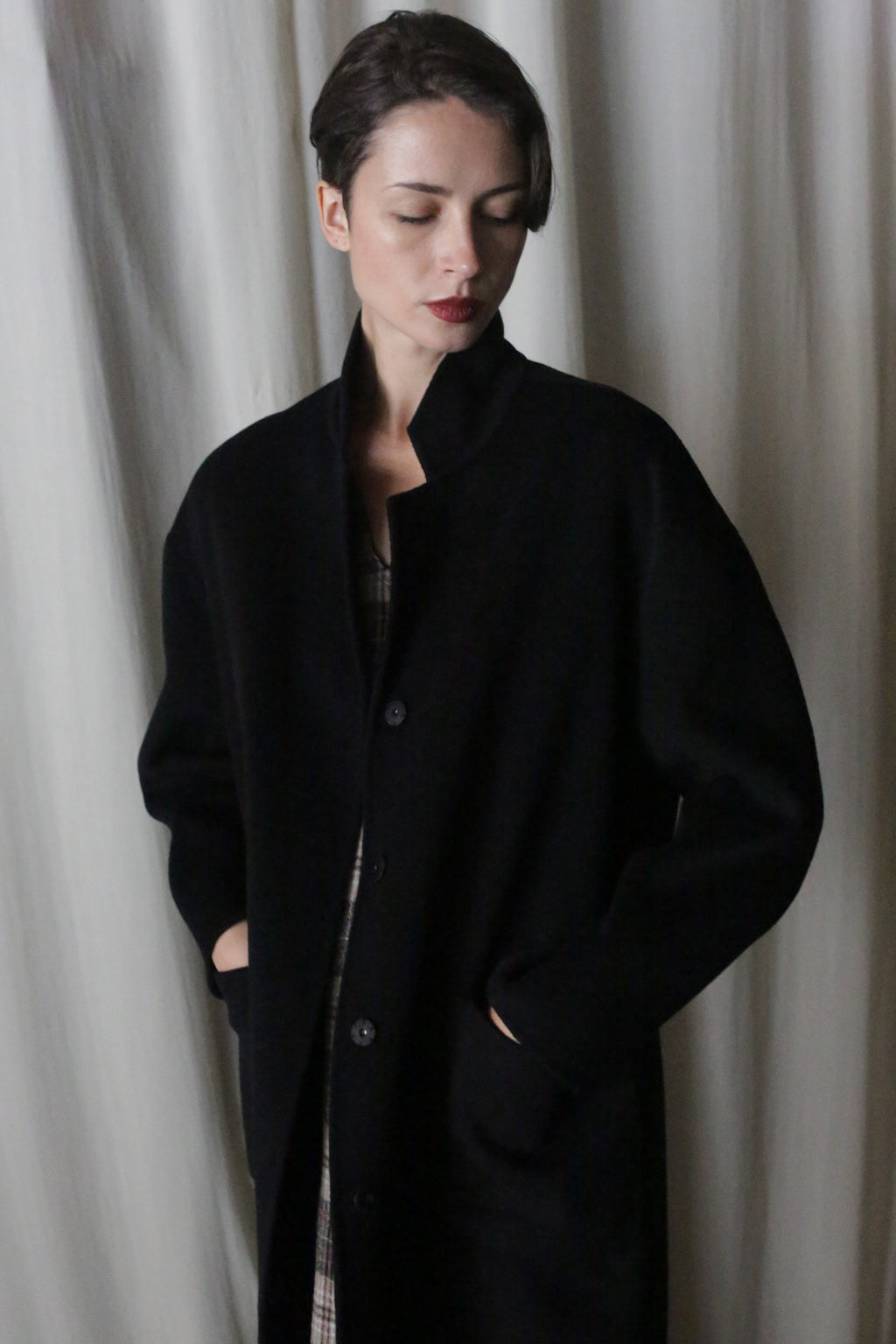 A person with short dark hair stands against a softly lit, light-colored curtain, hands in pockets, wearing the Bamford Long Double-face Cashmere in Black over a plaid shirt and looking slightly downward.