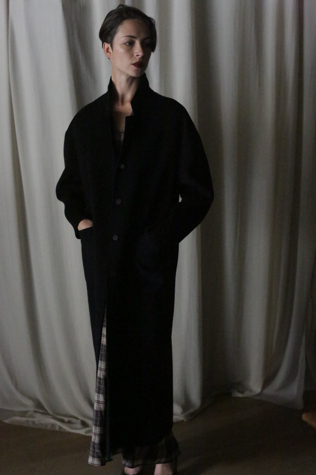 A person with short hair stands before a draped curtain, wearing the Bamford Long Double-face Cashmere coat in black with hands in pockets. A plaid garment peeks out beneath the coat under dim, moody lighting.