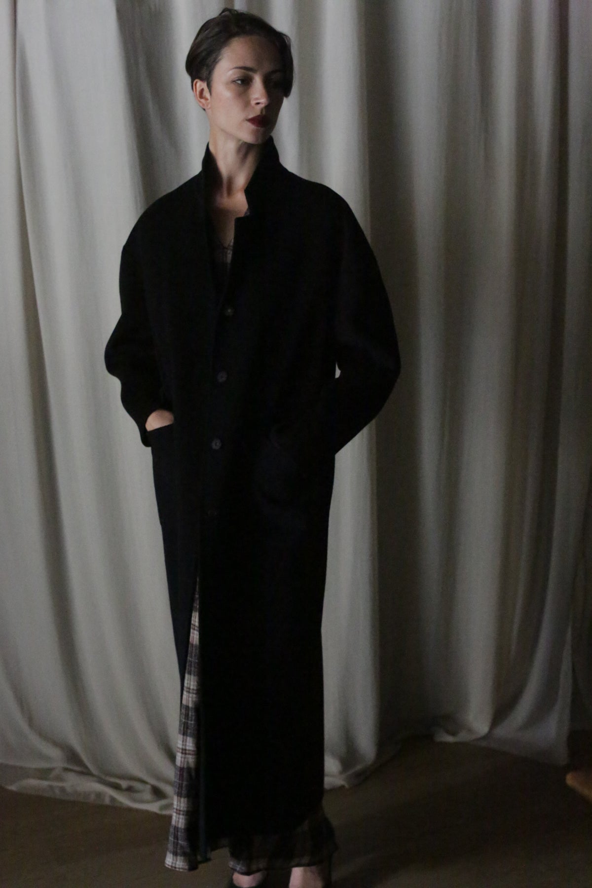 A person with short hair stands before a draped curtain, wearing the Bamford Long Double-face Cashmere coat in black with hands in pockets. A plaid garment peeks out beneath the coat under dim, moody lighting.