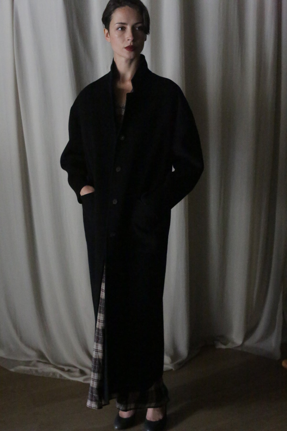 A woman stands indoors before cream curtains, wearing the Bamford Long Double-face Cashmere coat in black with her hands in the pockets. Plaid fabric peeks from beneath her outfit, and she finishes the look with black shoes.