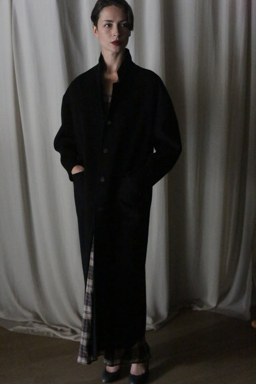 A woman stands indoors before cream curtains, wearing the Bamford Long Double-face Cashmere coat in black with her hands in the pockets. Plaid fabric peeks from beneath her outfit, and she finishes the look with black shoes.