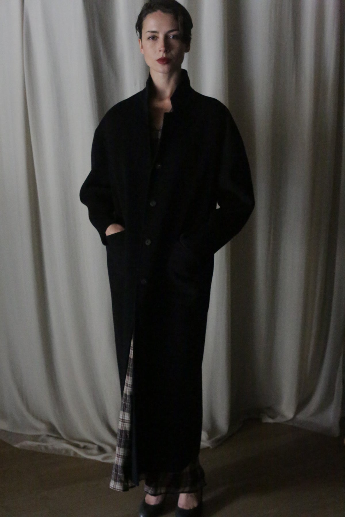 Indoors before cream curtains, a person wears the Bamford Long Double-face Cashmere coat in black with handmade snap buttons and hands in pockets, plaid pants peeking below. Lighting is dim and soft.