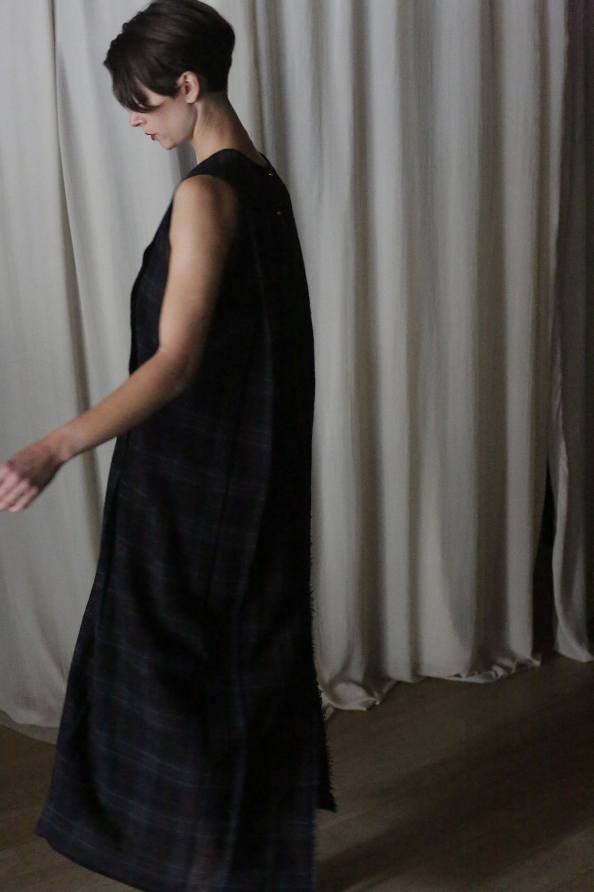 A person with short hair wearing The Winter Dress in Japanese Wool Gauze Forest Plaid exudes cozy sophistication as they walk by tall, white curtains in dim indoor lighting, facing sideways and moving gracefully to the left.