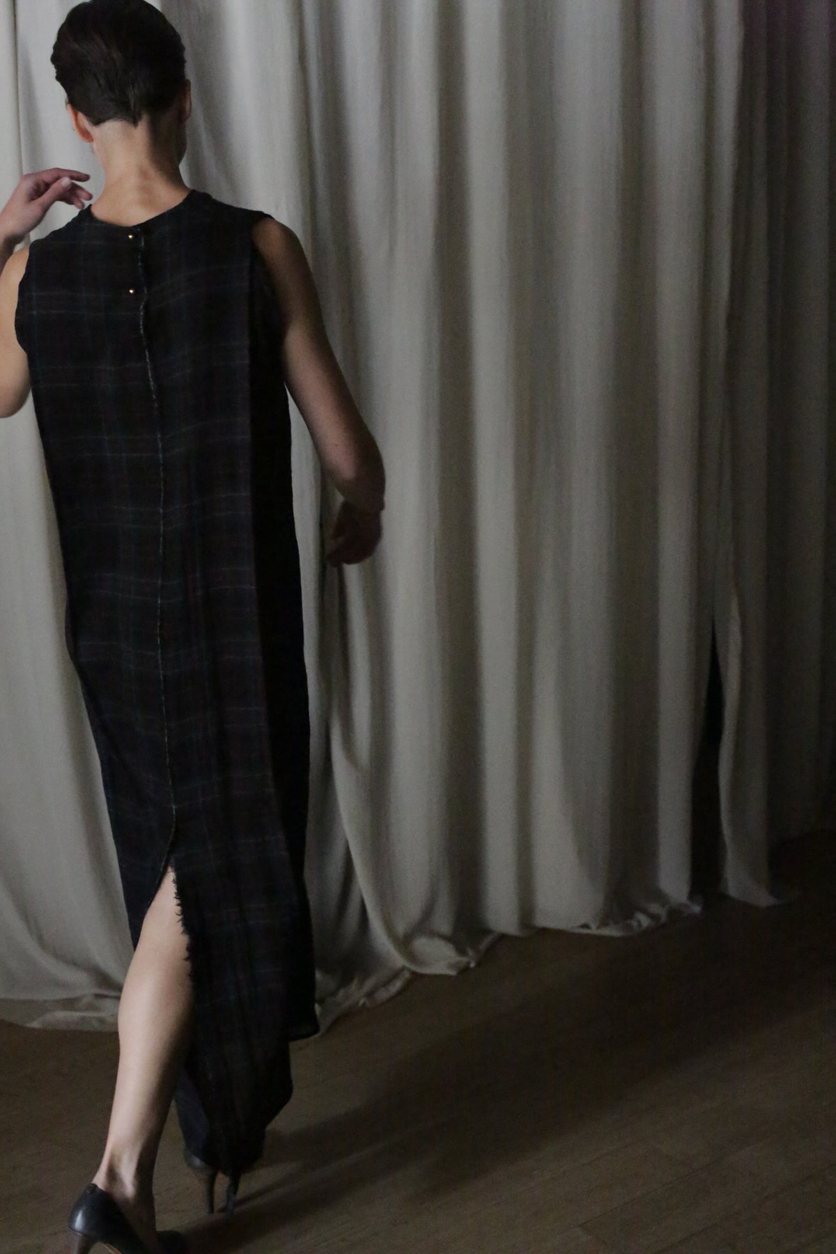 Wearing "The Winter Dress" in Japanese Wool Gauze Forest Plaid, a person with short hair moves through a dimly lit room. The sleeveless, floor-length black dress with its subtle plaid pattern and back slit adds cozy sophistication against beige curtains.
