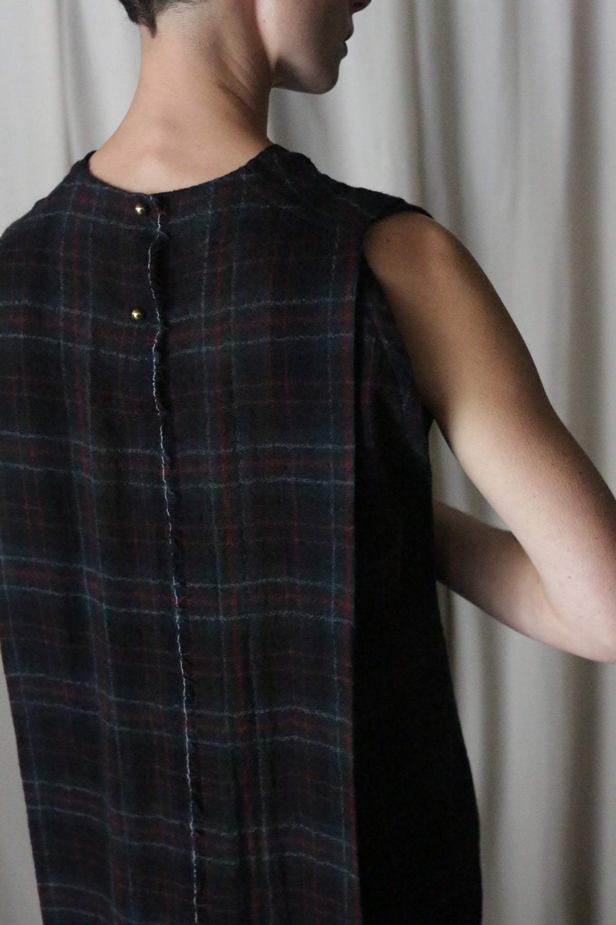 A person wearing "The Winter Dress" in Japanese Wool Gauze Forest Plaid, a sleeveless garment, stands side-on to the camera. Their short hair is visible as their head turns away against a light-colored curtain backdrop.