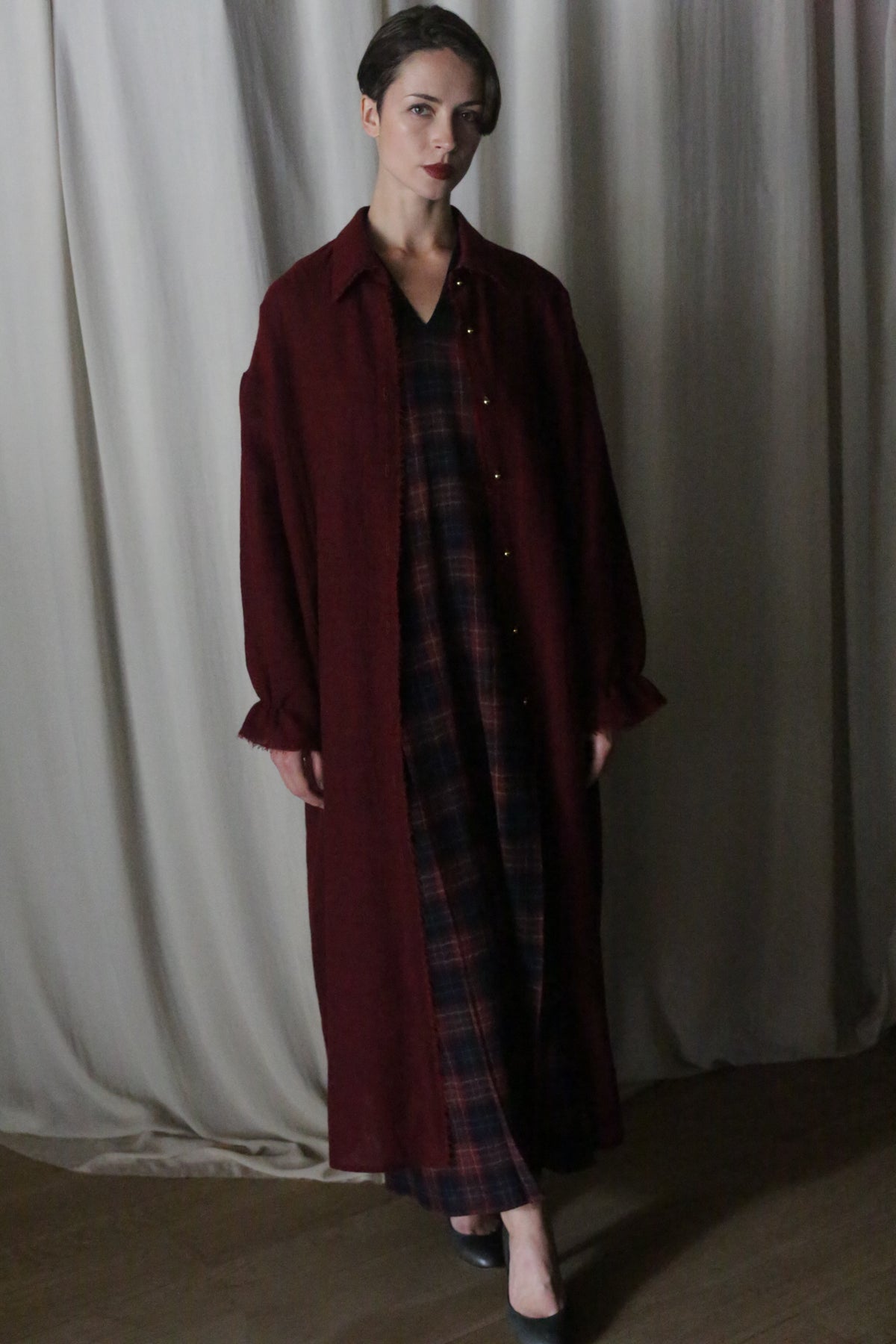 A woman indoors stands before light curtains, wearing The Winter Dress in Japanese Wool Gauze Wine Plaid under a long burgundy coat and black shoes. She has short hair and a calm expression.