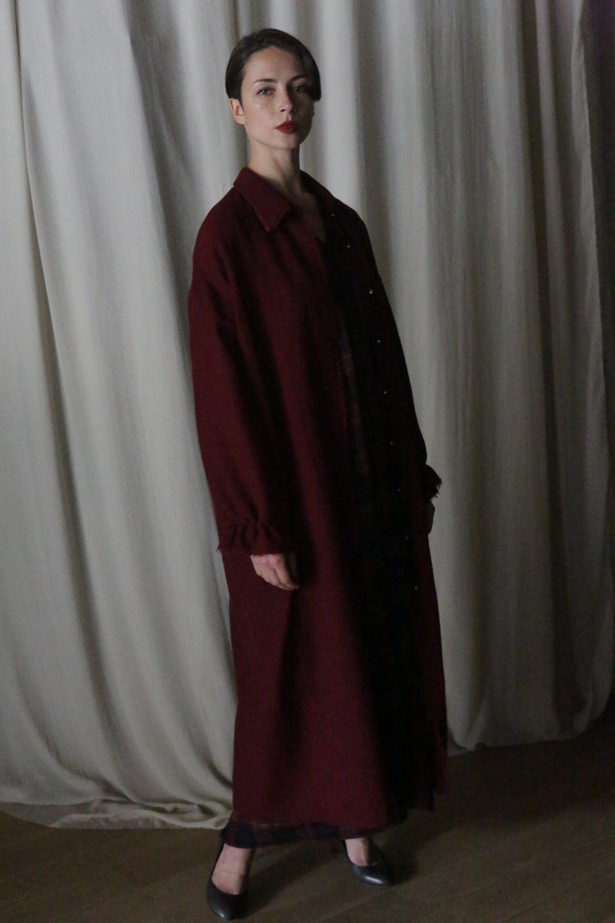 A woman with short hair wears the Dress Coat | Japanese Wool Gauze Wine and black shoes, standing before cream-colored curtains and looking at the camera with a neutral expression.