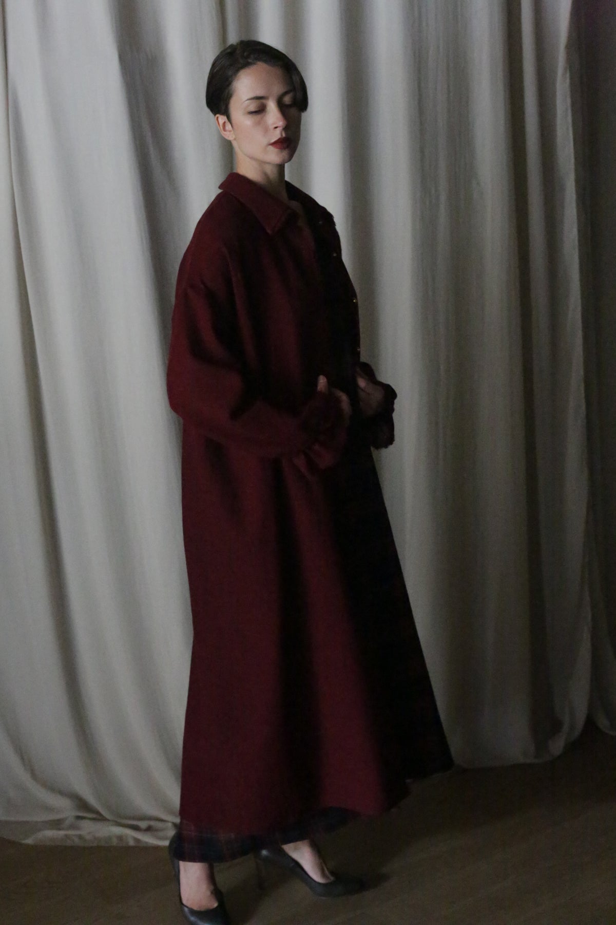A person with short dark hair stands indoors in front of cream curtains, wearing the Dress Coat | Japanese Wool Gauze Wine—a long, loose-fit maroon coat—enveloped in soft, dim lighting.